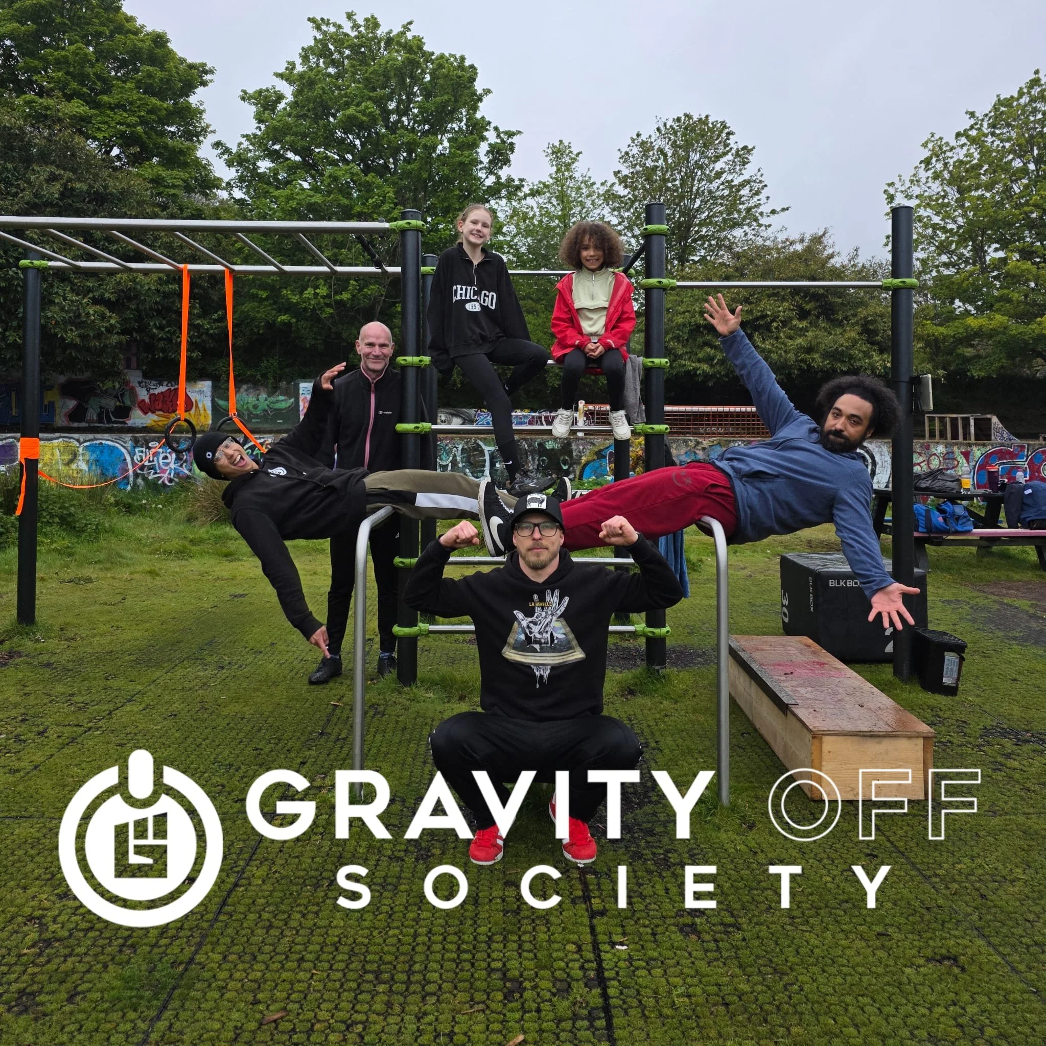 Group of five people outdoors, three men and two women, posing on a playground structure with graffiti in the background. Two women are sitting on top, one man and another woman are sitting on the ground, and the fifth person is lying across the stru