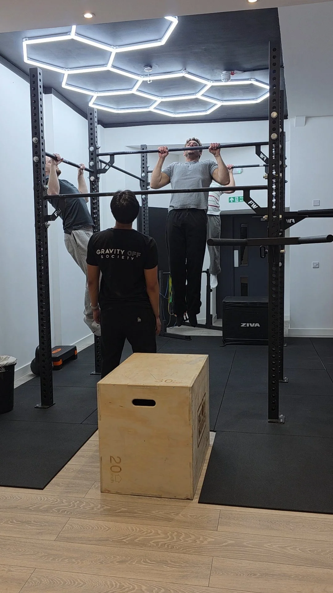 Three men working out in a gym, two doing pull-ups on a black pull-up bar, one supervising, with a wooden plyometric box in the foreground and a hexagonal LED ceiling light.