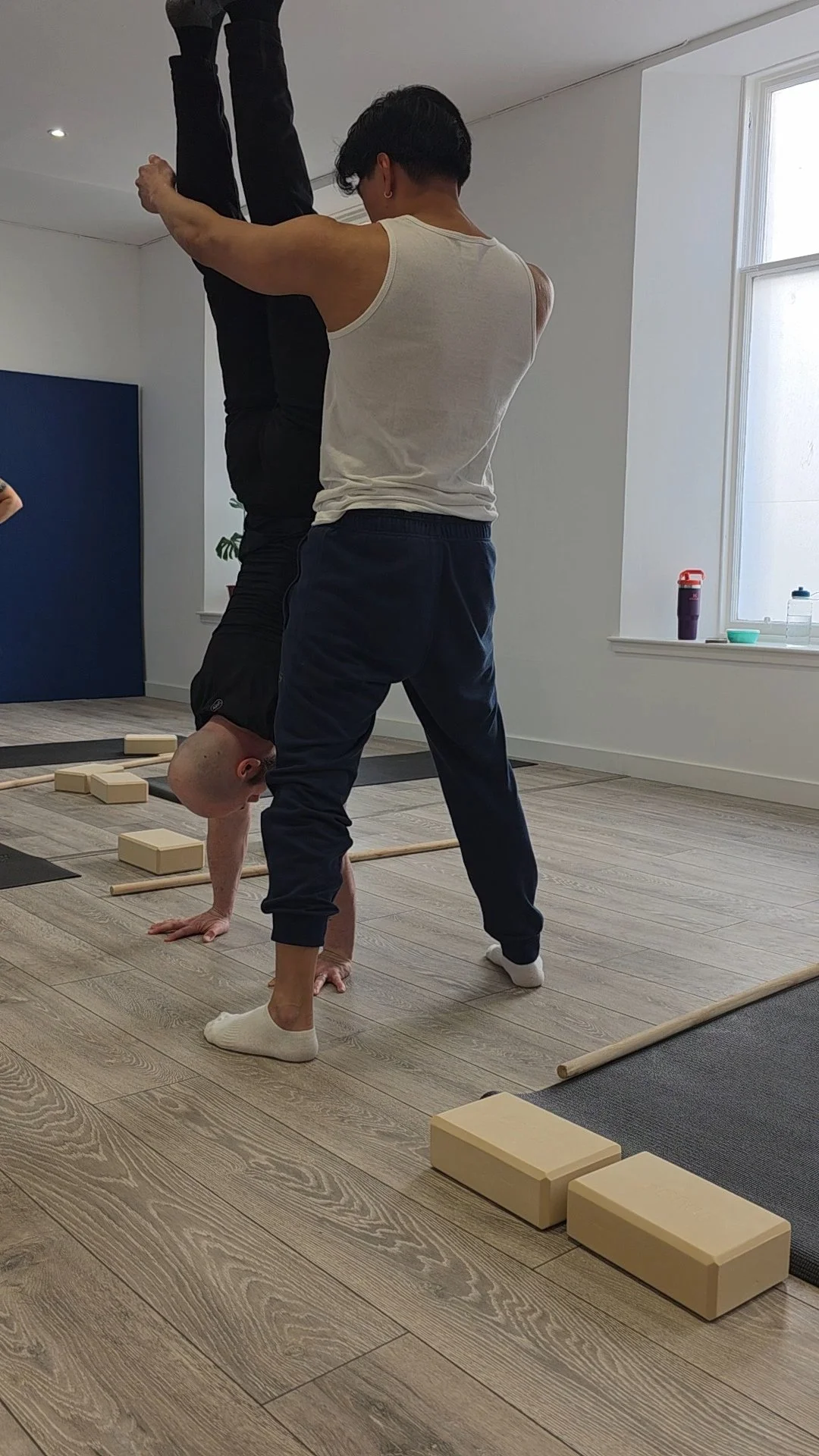 Two men practicing yoga or acrobatics in a brightly lit indoor space with wooden flooring, yoga blocks, and mats.