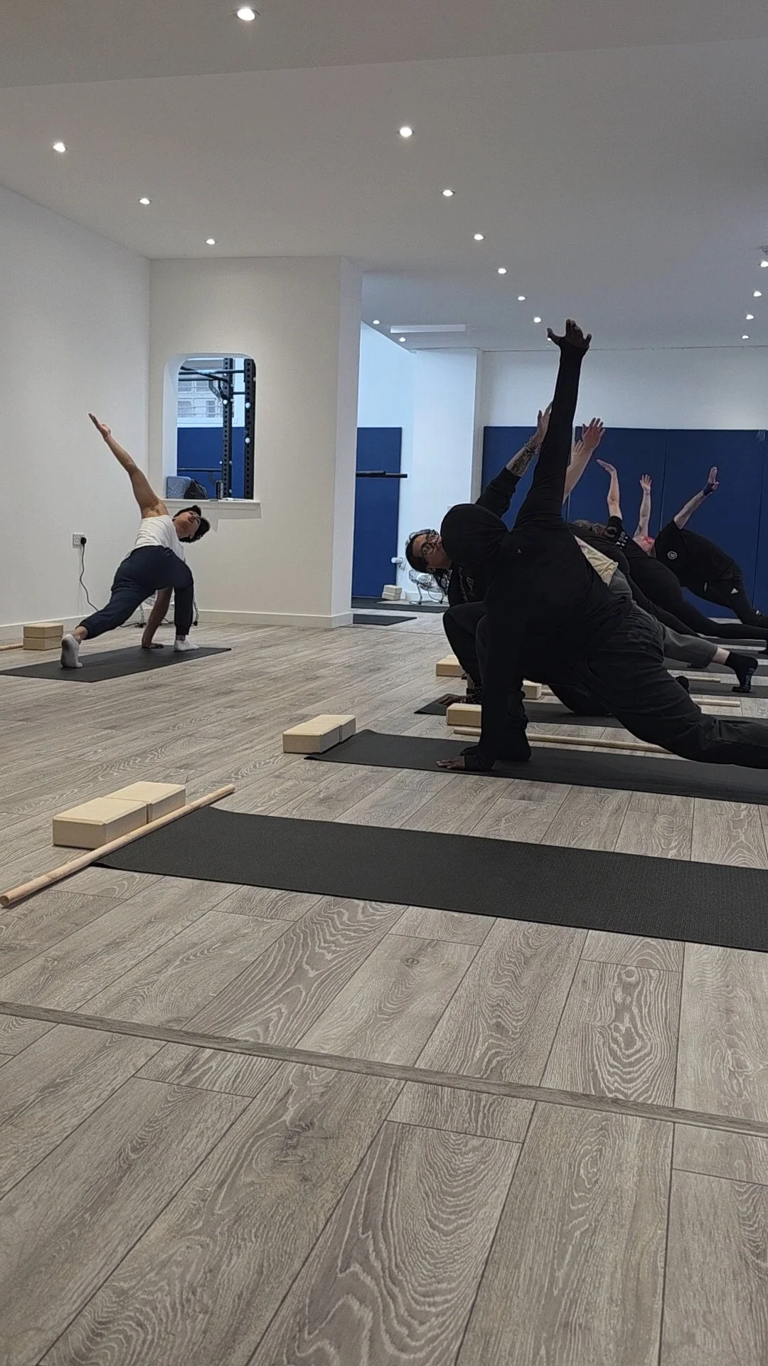 A group of people practicing yoga in a studio with wooden flooring, black mats, and yoga blocks. One person in white is leading, while others in black are following in side stretches.