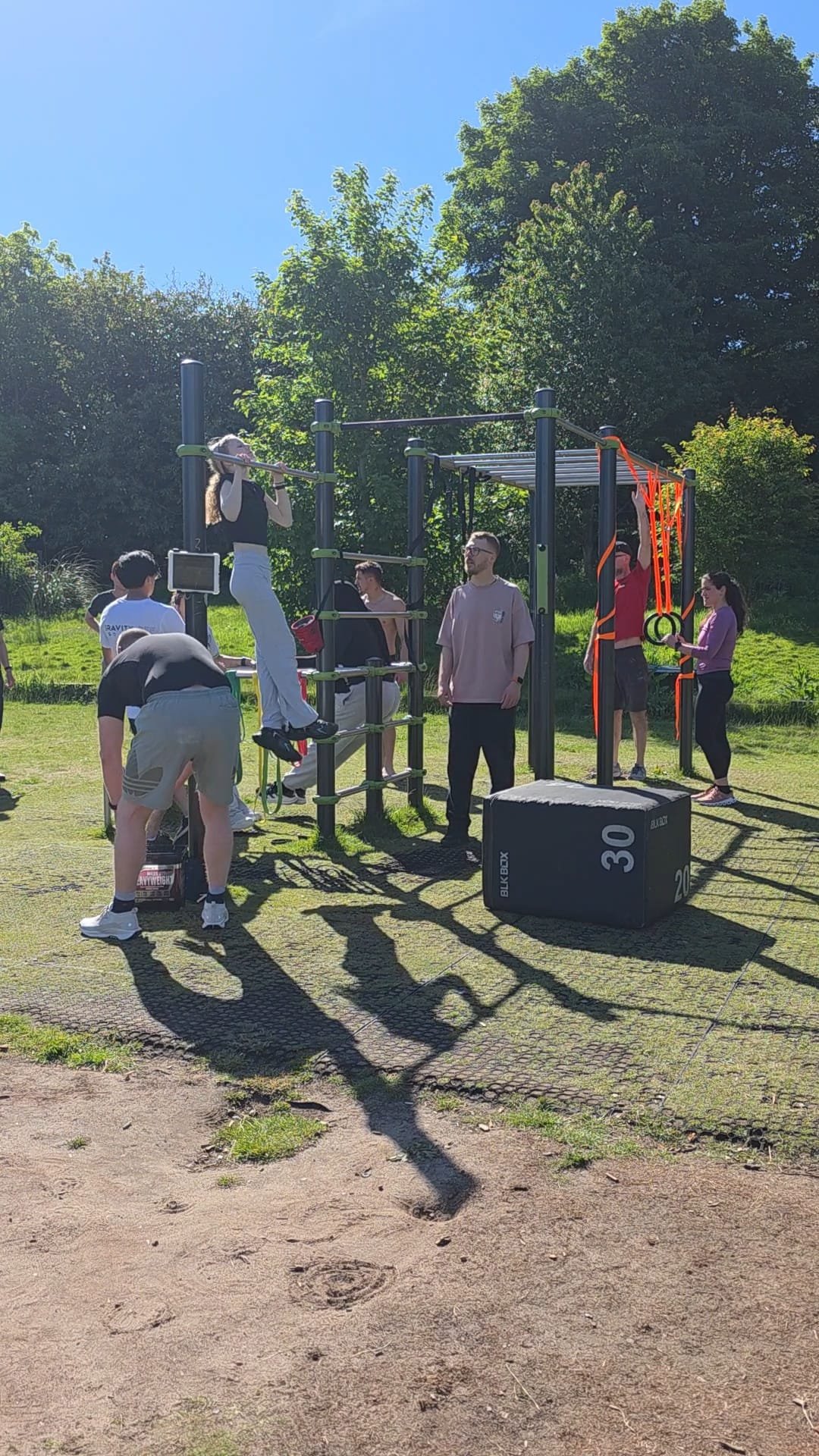 People exercising outdoors on fitness equipment, including pull-up bars and suspension straps, with trees and a blue sky in the background.