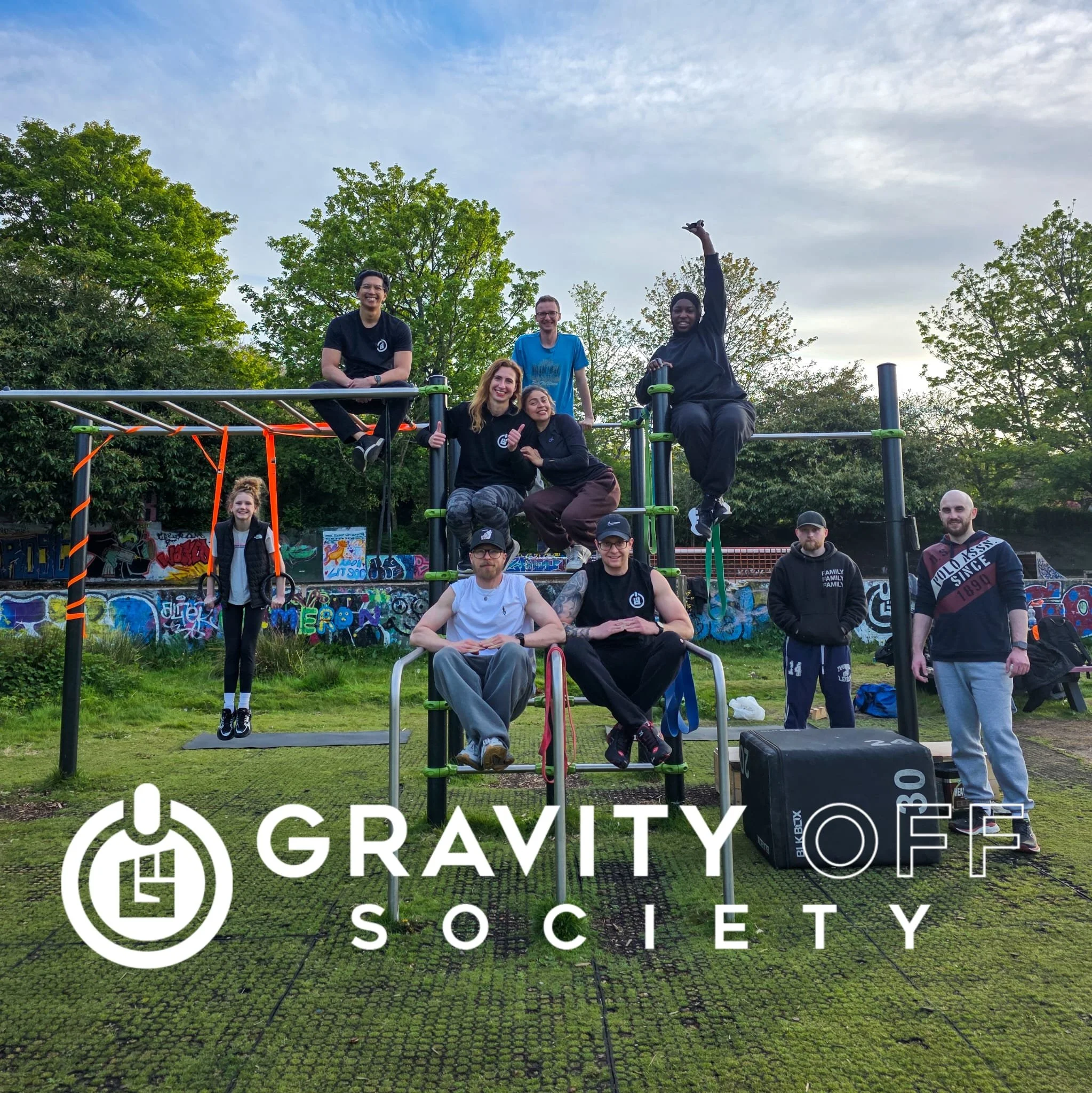 Group of smiling people at an outdoor park or skatepark with graffiti on the walls and trees in the background. Some are sitting, others standing, one person is raising an arm, and a few are on skateboards.