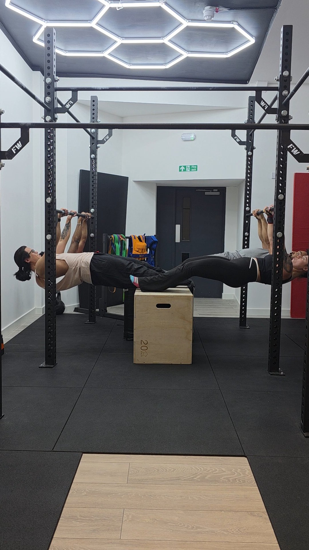 Two women are exercising in a gym, lying face down on a bench with their legs extended and holding a barbell above their shoulders. One woman is on the left and the other is on the right, with a wooden plyometric box between them.