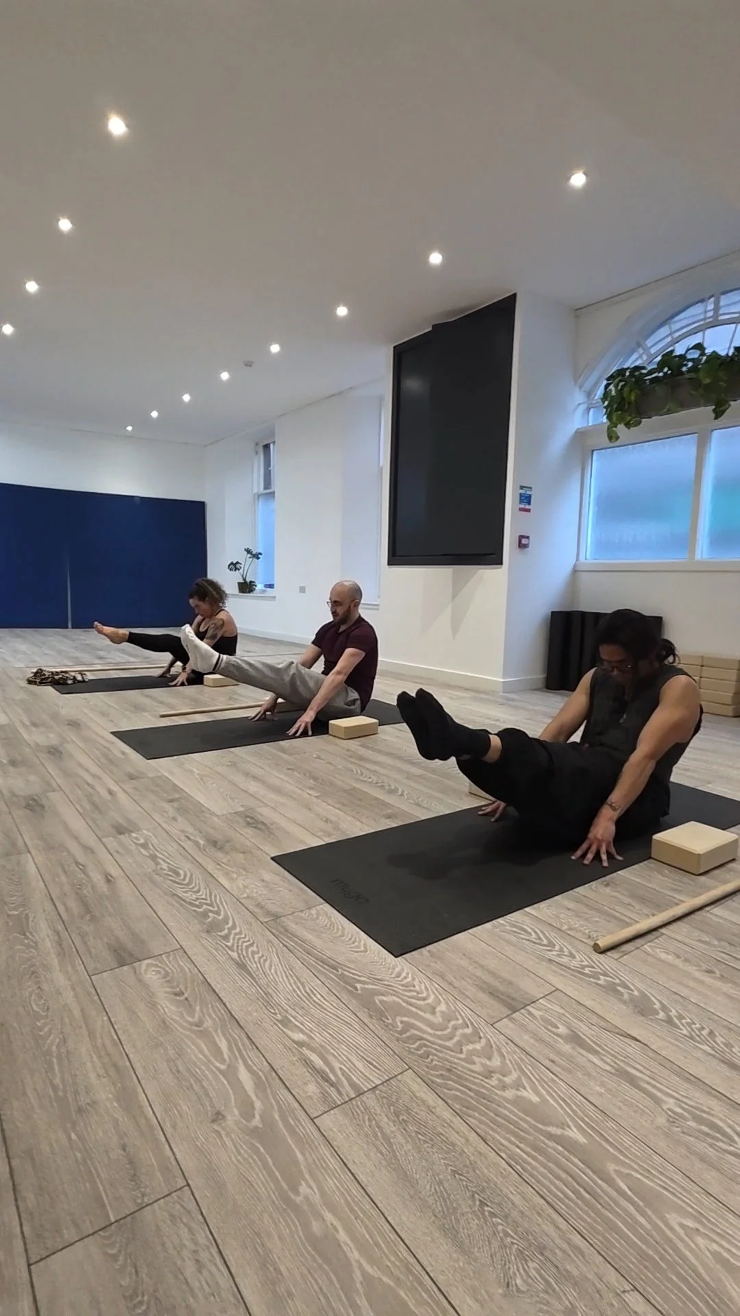 Three people participating in a yoga class in a studio, practicing side stretches on yoga mats, with blocks and straps, in a well-lit room with wooden floors and white walls.