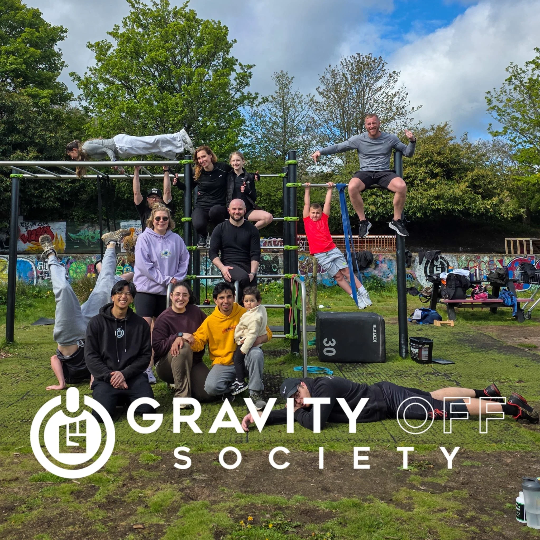 Group of people at an outdoor fitness event with a climbing structure and graffiti mural in the background, organized by Gravity Society.