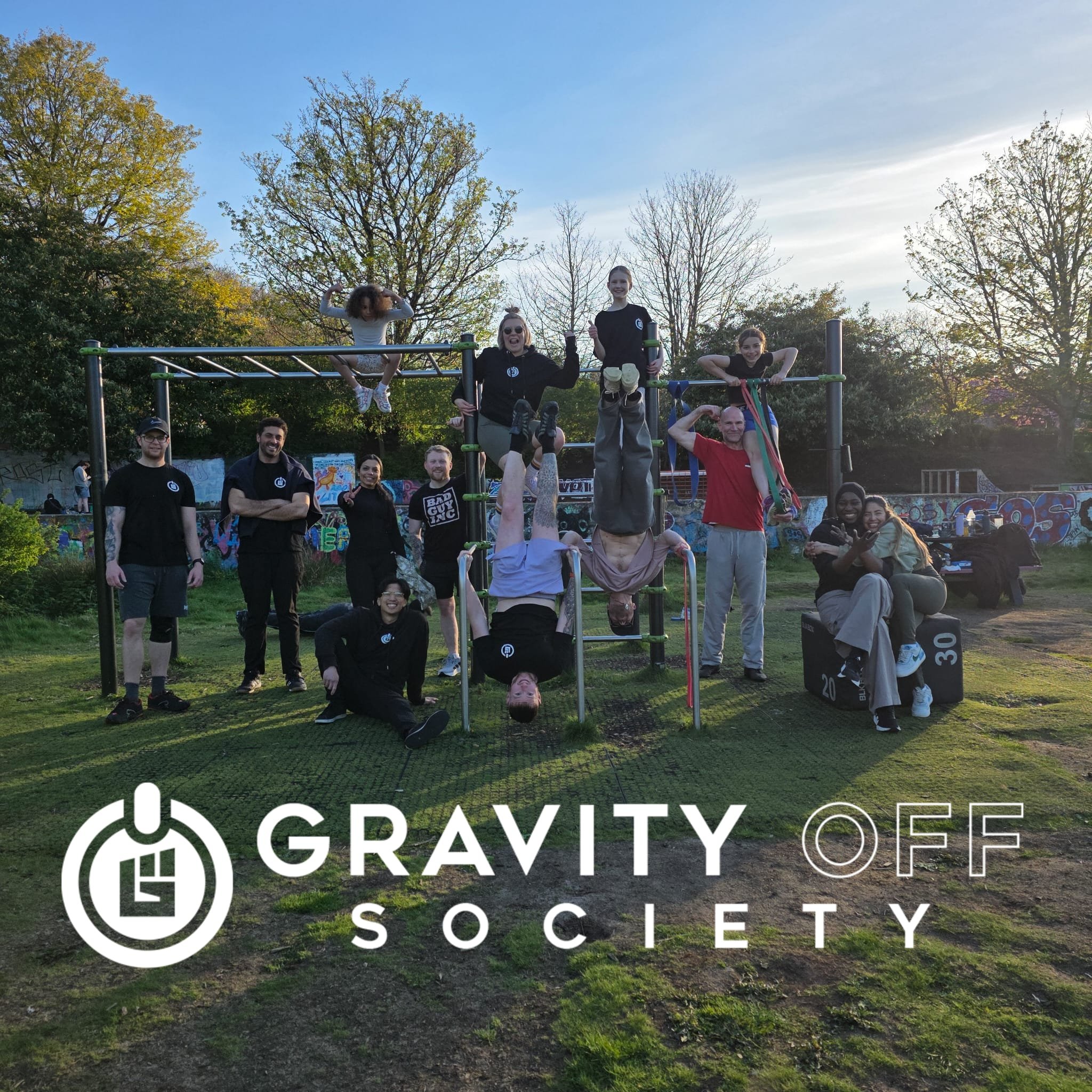 Group of people at an outdoor event, some climbing a playground structure, with a graffiti-covered wall in the background and a large logo for the Gravity Society at the bottom of the image.