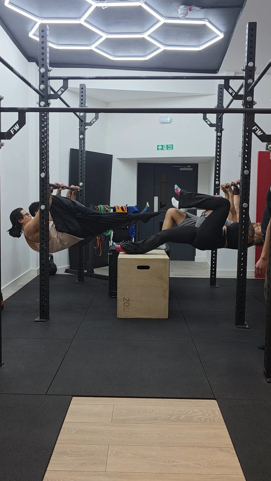 Two people performing a suspension workout on pull-up bars inside a gym, with a wooden plyometric box and LED ceiling lights.