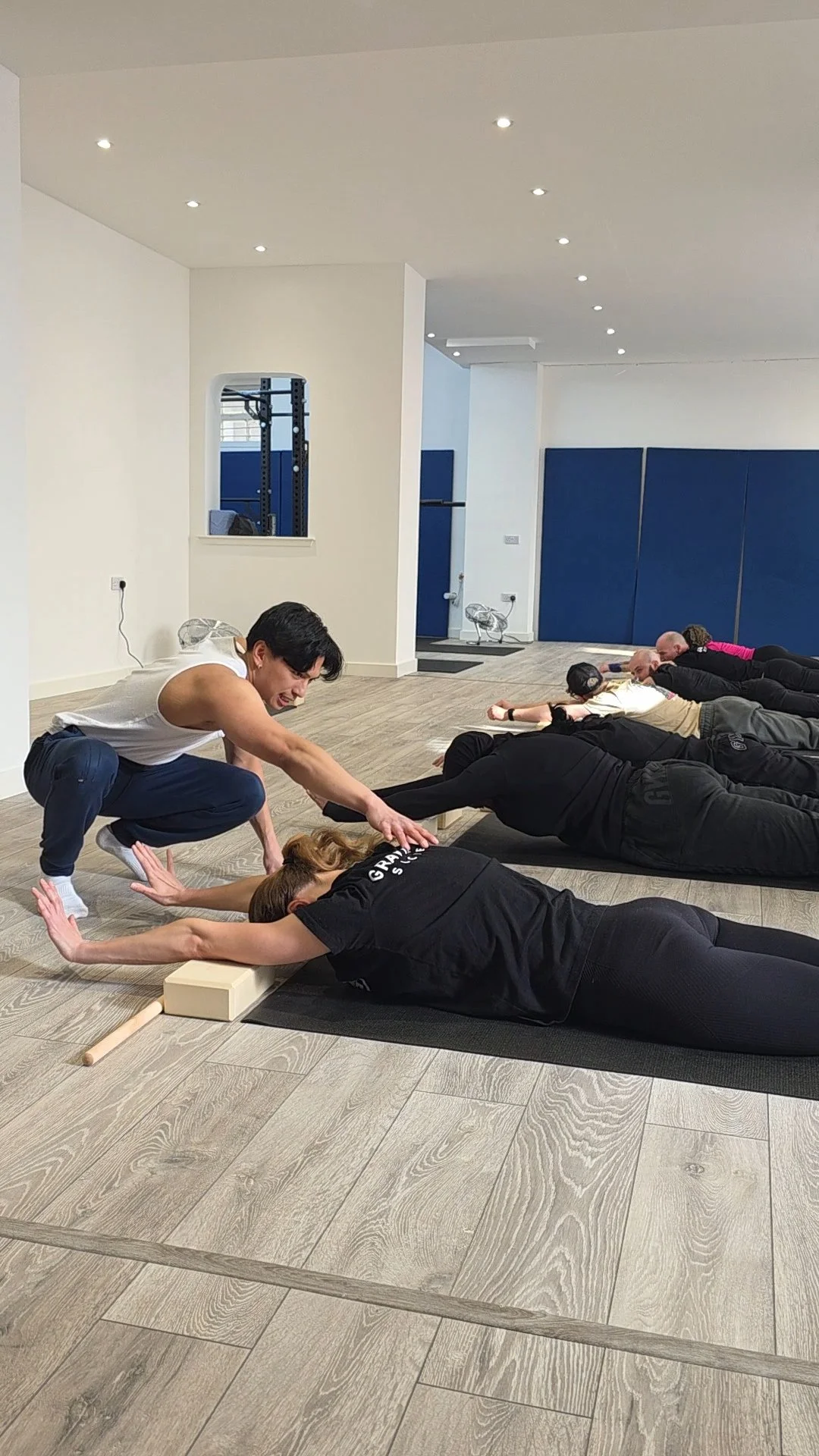 A yoga class in a studio with participants lying on their stomachs in a pose, receiving instruction from an instructor.