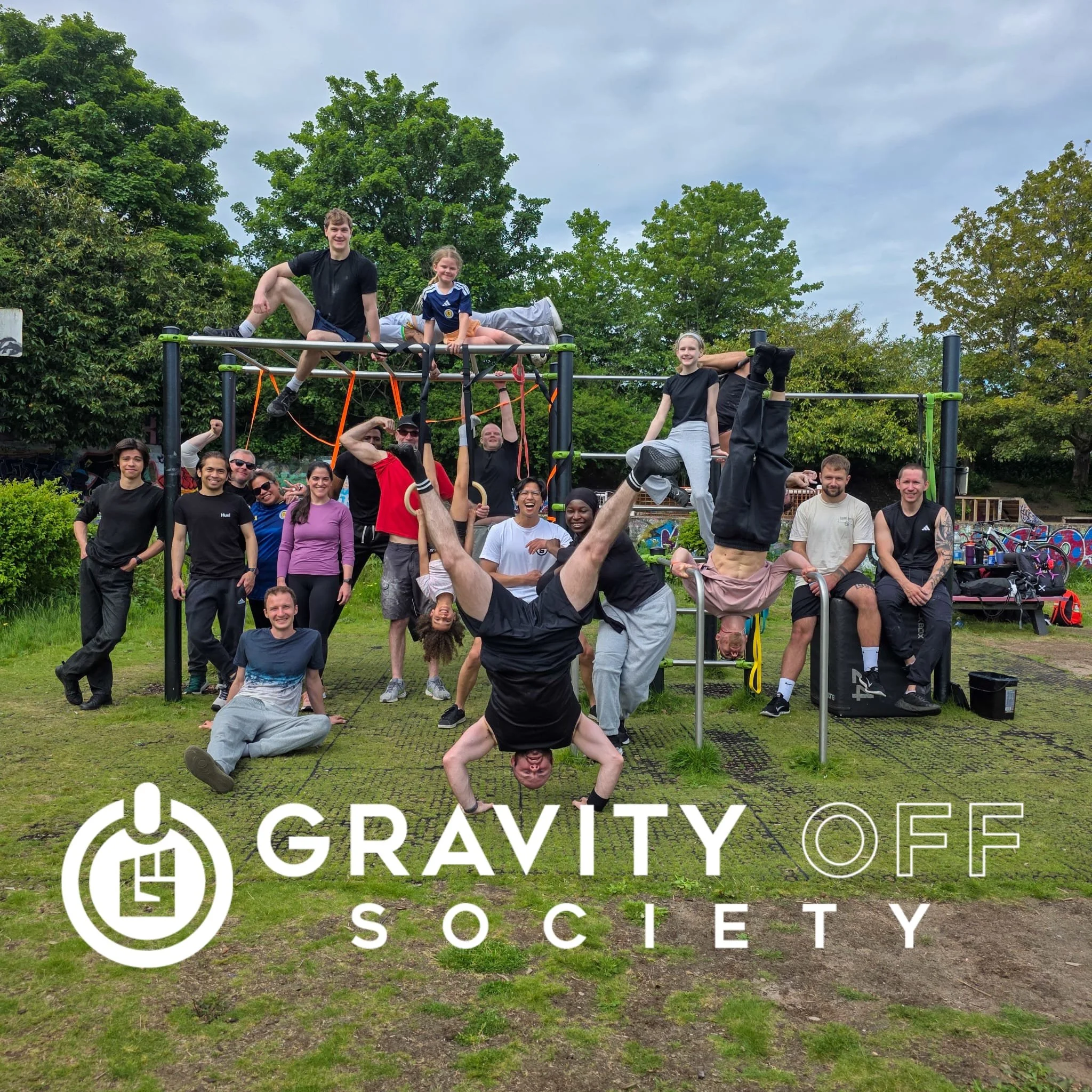 Group of people gathered outdoors in front of a climbing structure, some sitting and others standing, with trees and cloudy sky in the background, participating in a community event organized by Gravity Society.