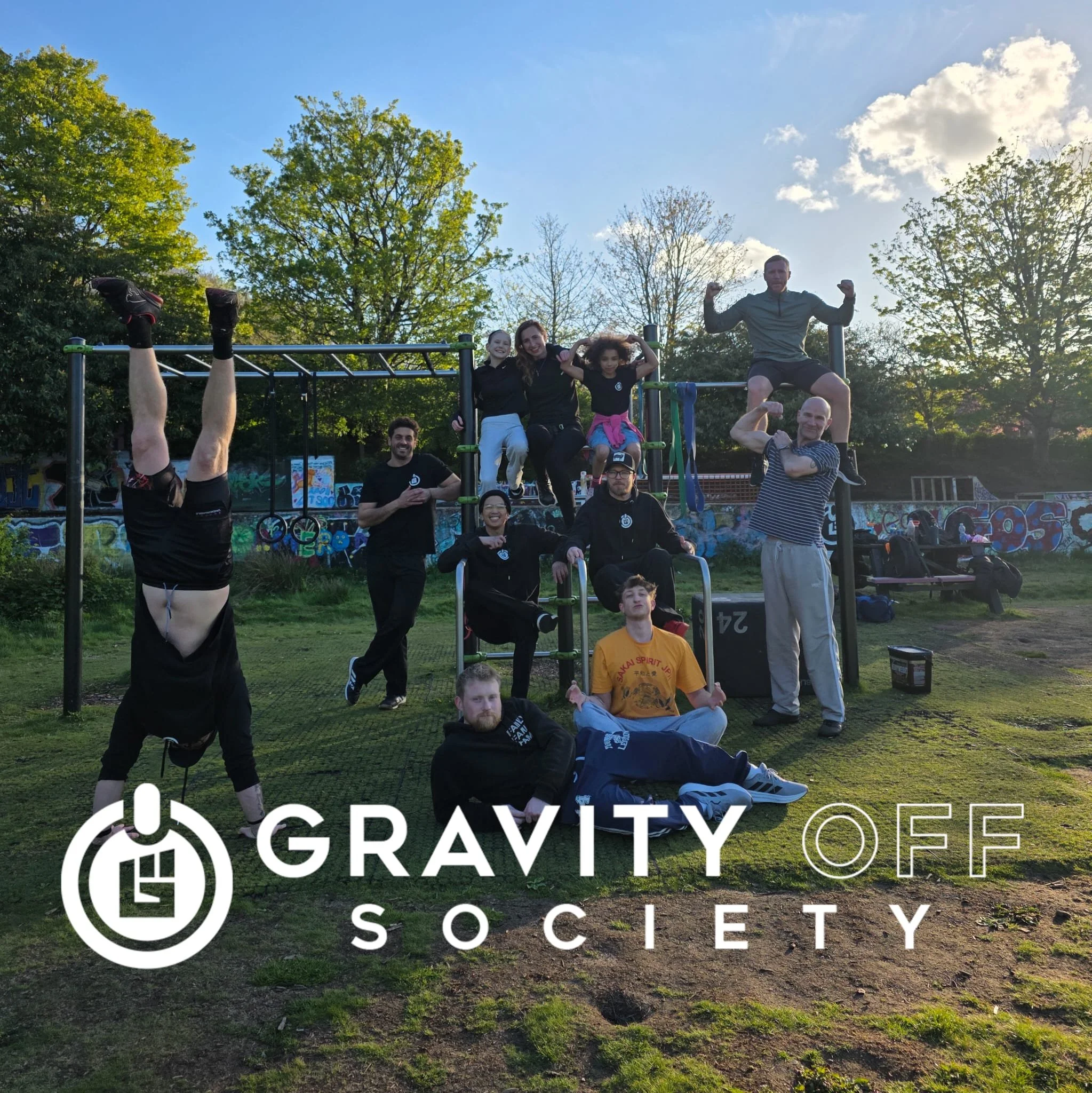 A group of people at a park outdoor fitness area with trees and a bright blue sky. Some are sitting, flexing arms, or hanging upside down on playground equipment. The image has a logo and text for 'Gravity Society Societ'.