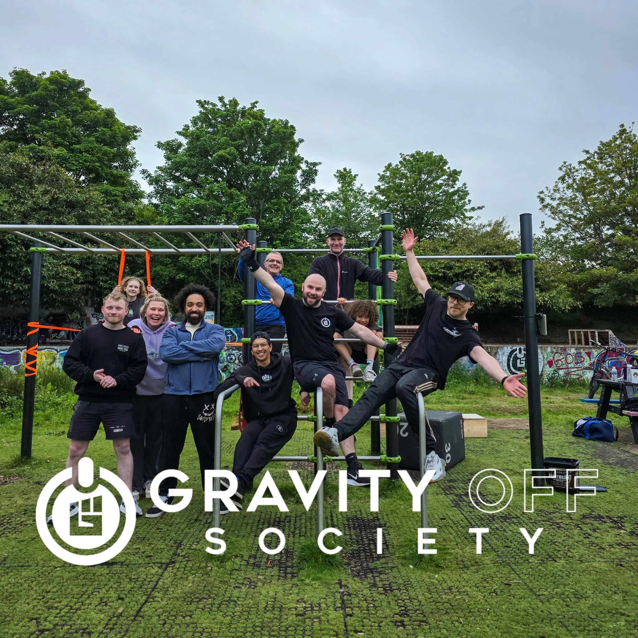 Group of people standing and sitting outdoors in front of a fitness structure, smiling and waving, with trees and graffiti wall in the background, at Gravity Society gym.