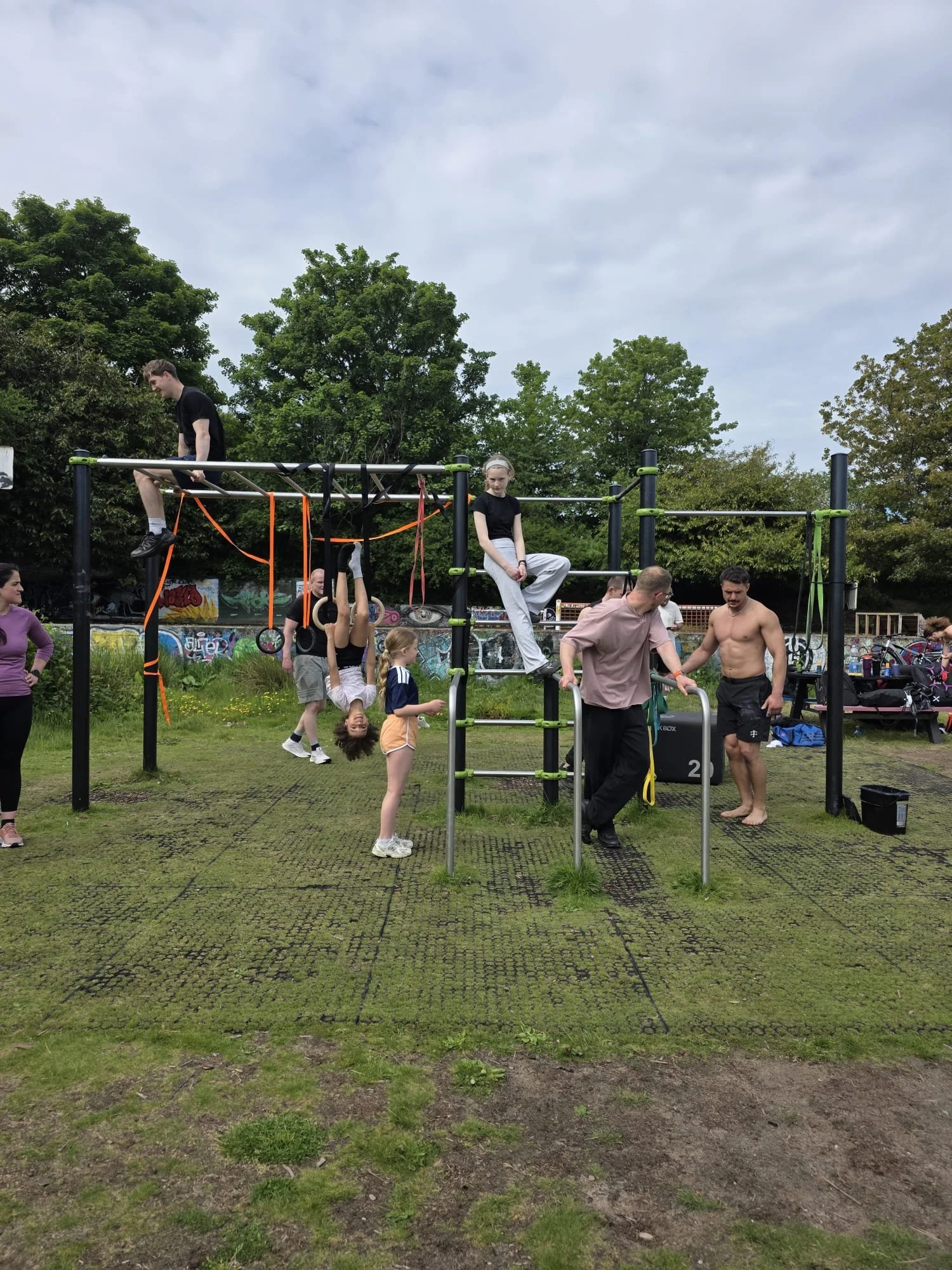 People exercising outdoors on a green moss-covered surface with trees and graffiti in the background. Some are hanging from gymnastic rings, sitting on the equipment, or talking. A man is shirtless, and the sky is cloudy.