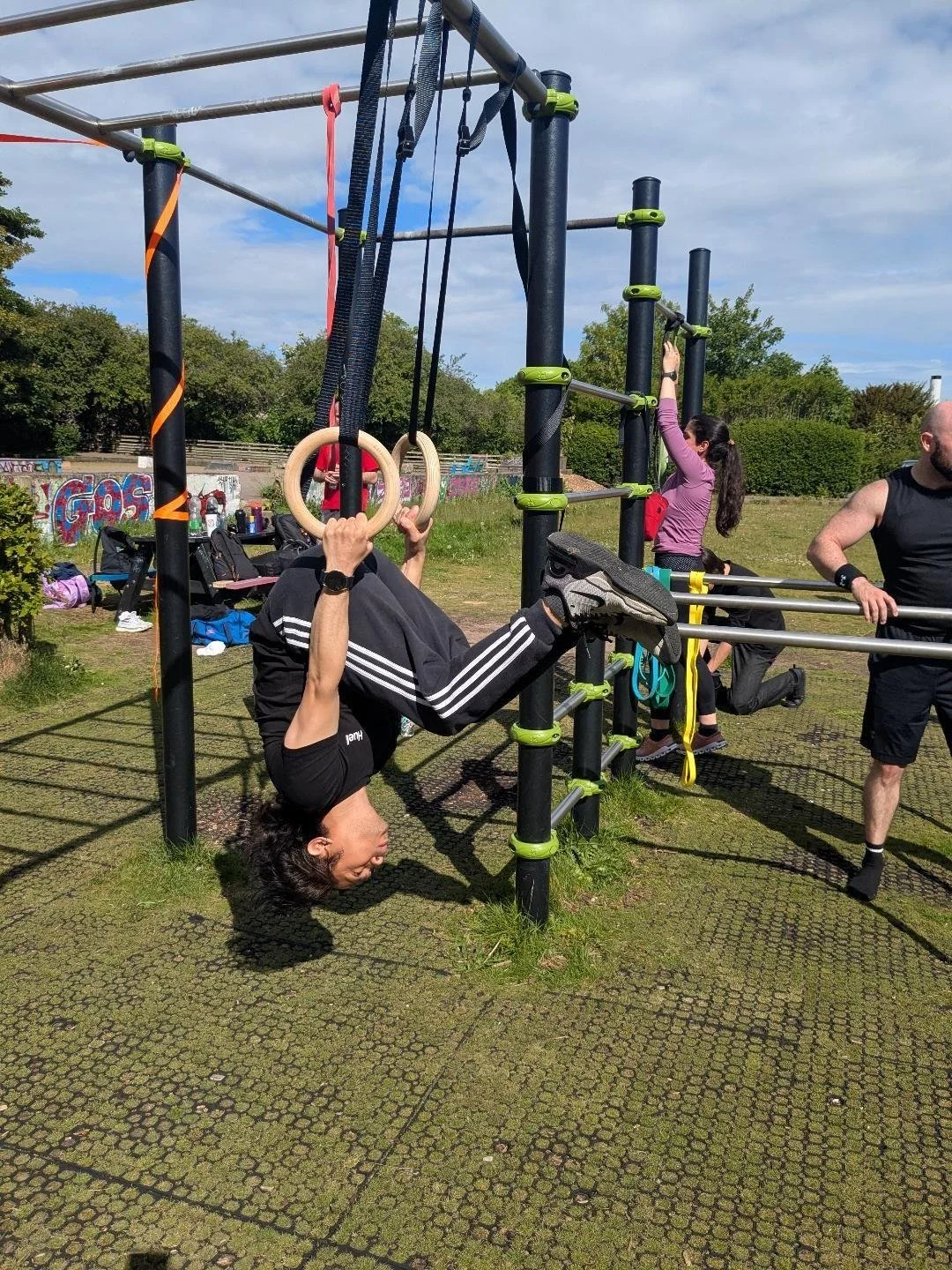 Person hanging upside down using gymnastic rings at an outdoor fitness park, with other individuals working out on horizontal bars in the background.