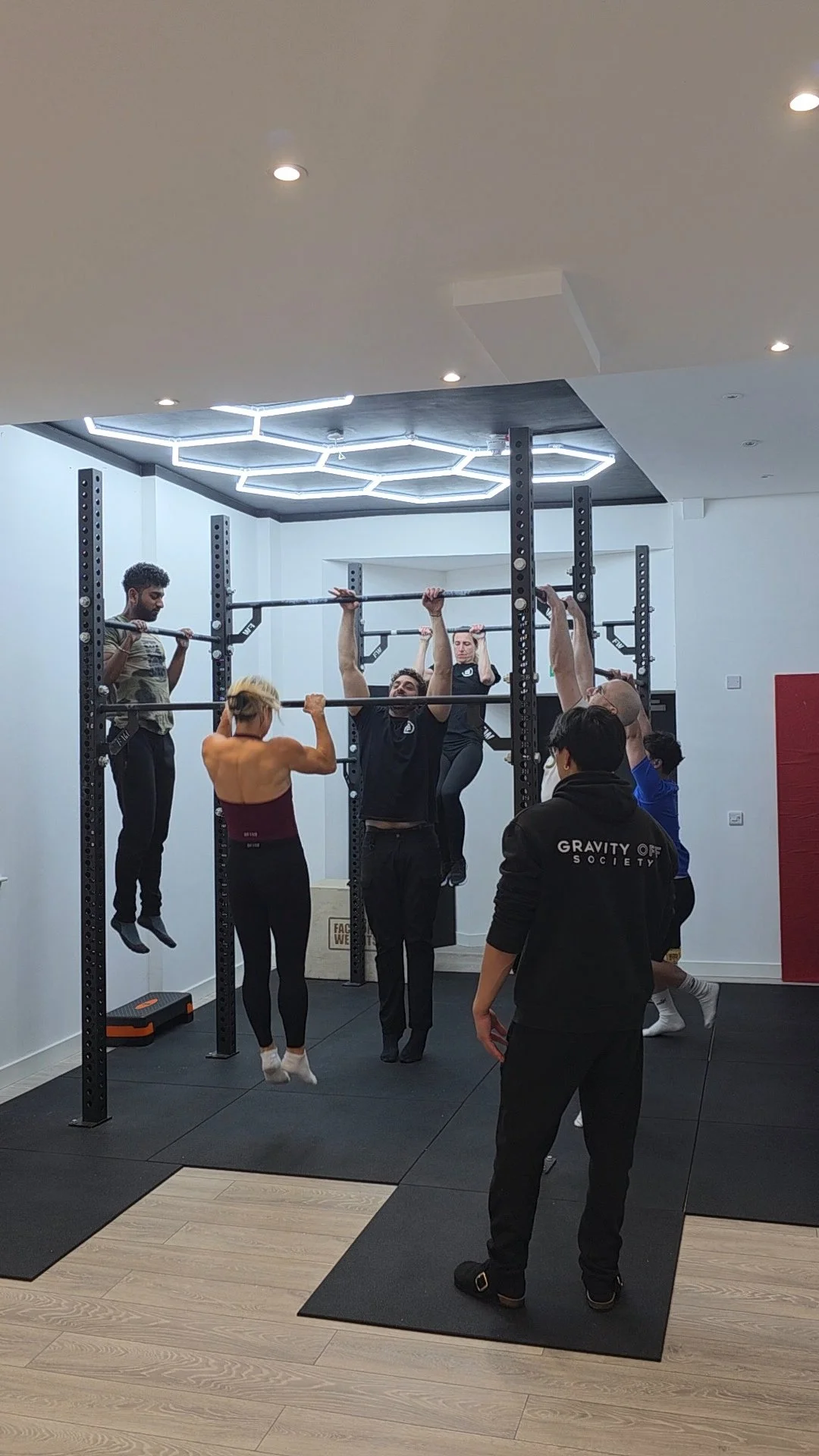 People training in a gym using pull-up bars, with some hanging and others assisting or observing, under modern lighting.