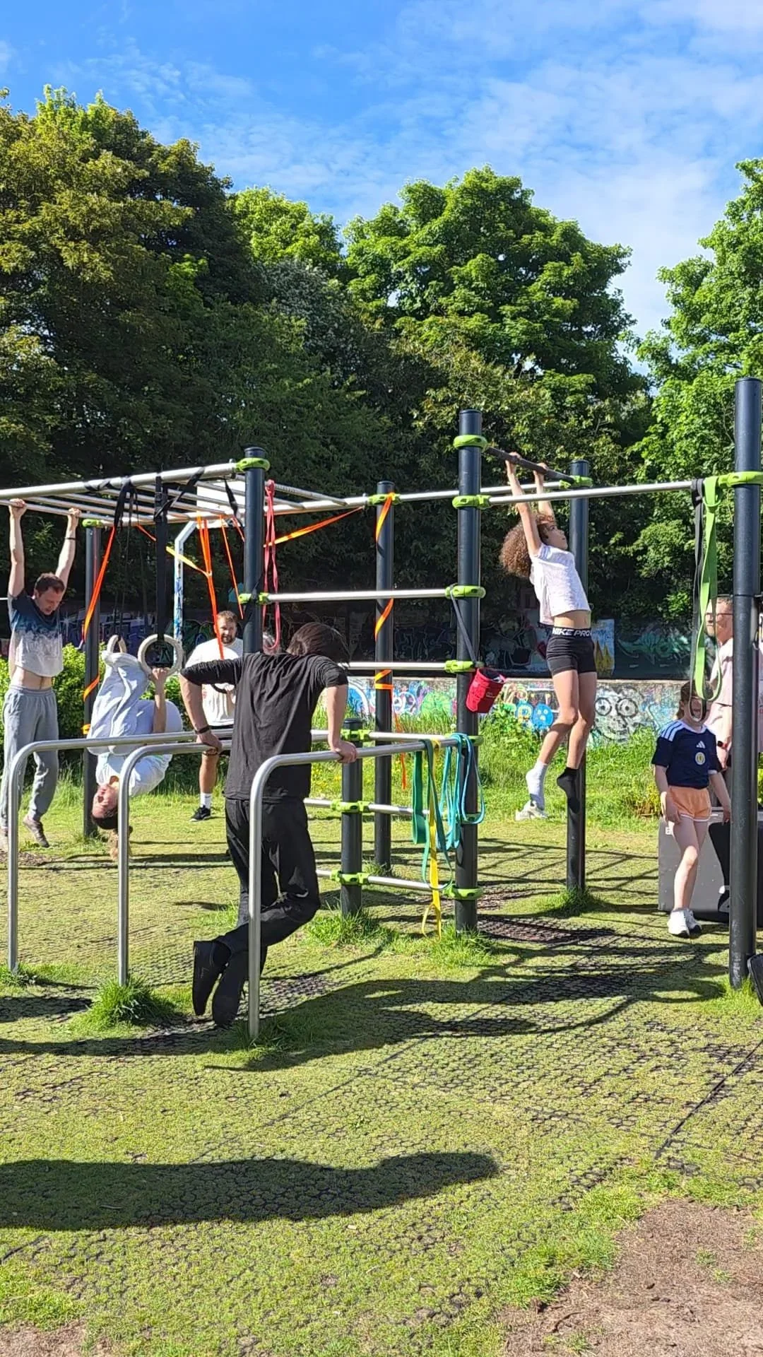 Children and adults playing on outdoor gym equipment in a park with green trees and a blue sky.