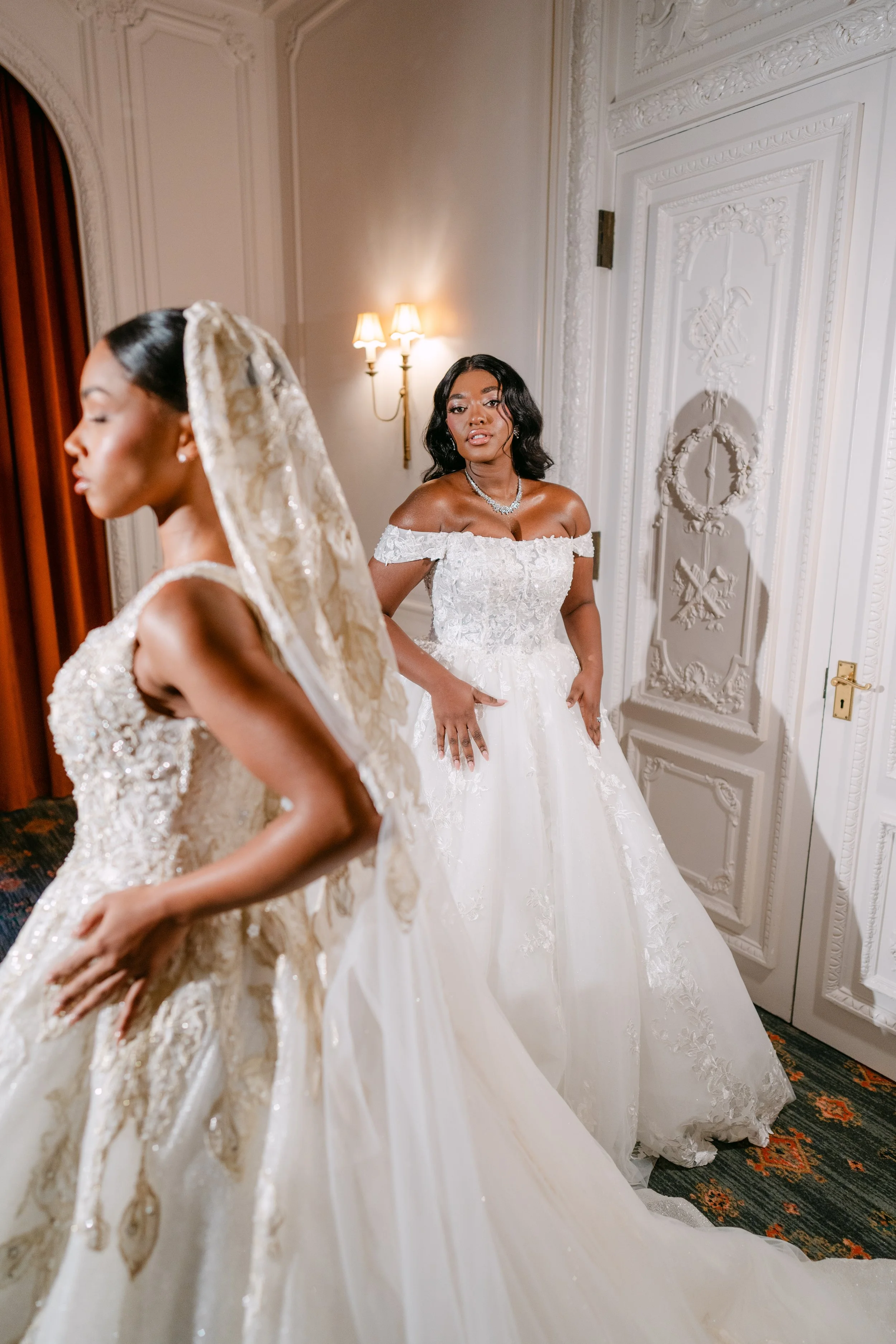 Two women in elegant wedding dresses, one with a lace veil, standing in a luxurious room with ornate white walls and a patterned carpet. One woman is in the foreground and the other in the background, both appearing to prepare for a wedding.
