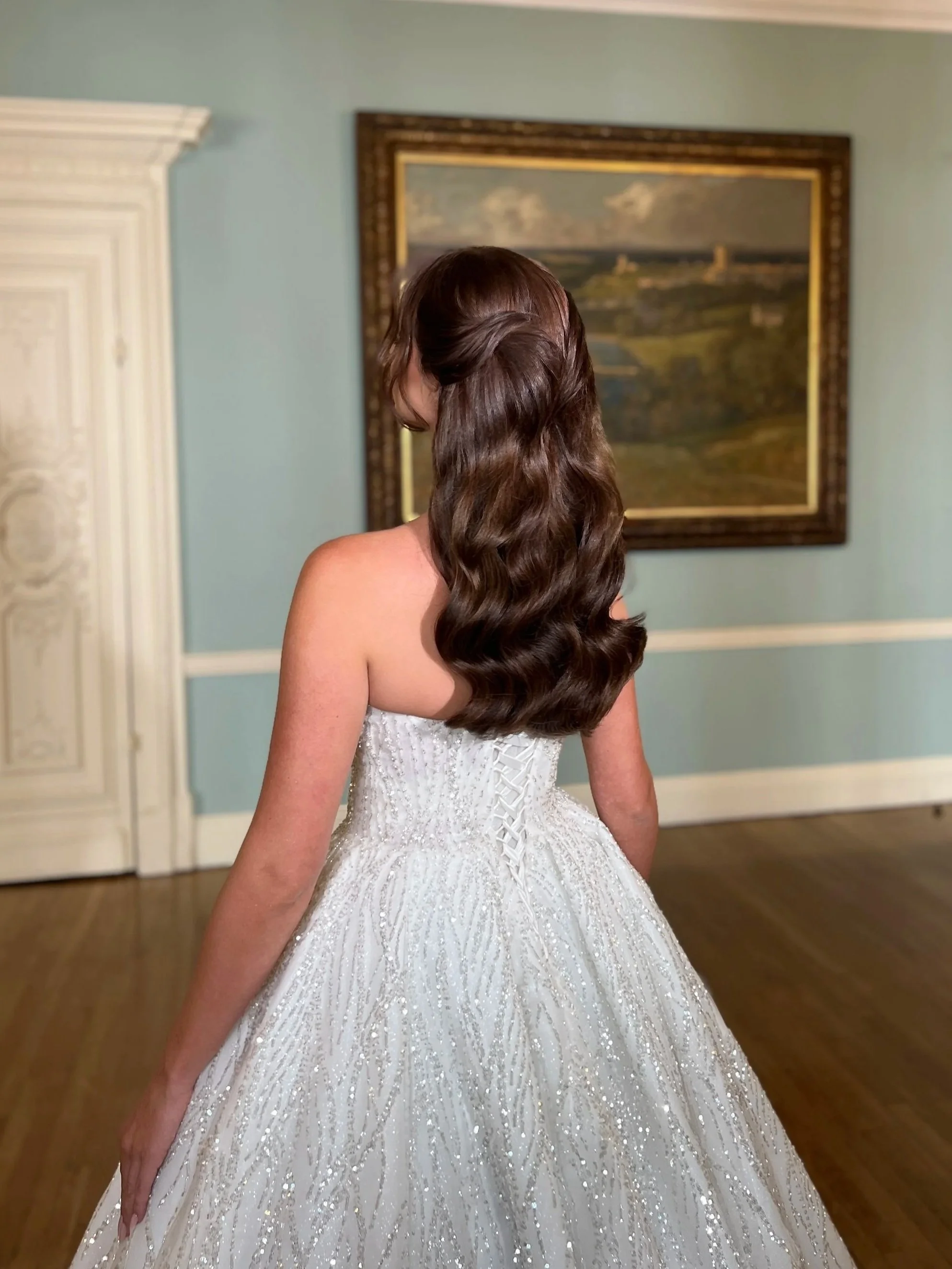 A bride with long, wavy brown hair in a strapless white evening gown with intricate beading and embroidery, standing in a room with light blue walls, a large framed landscape painting, and a white decorative fireplace.