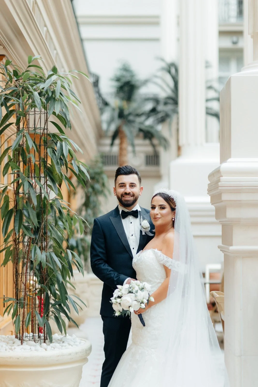 A bride and groom standing together inside a grand venue. The groom is wearing a black tuxedo with a bow tie, and the bride is in a white wedding gown with lace details, holding a bouquet of white flowers. They are smiling and posing for the camera near large white columns and lush greenery.