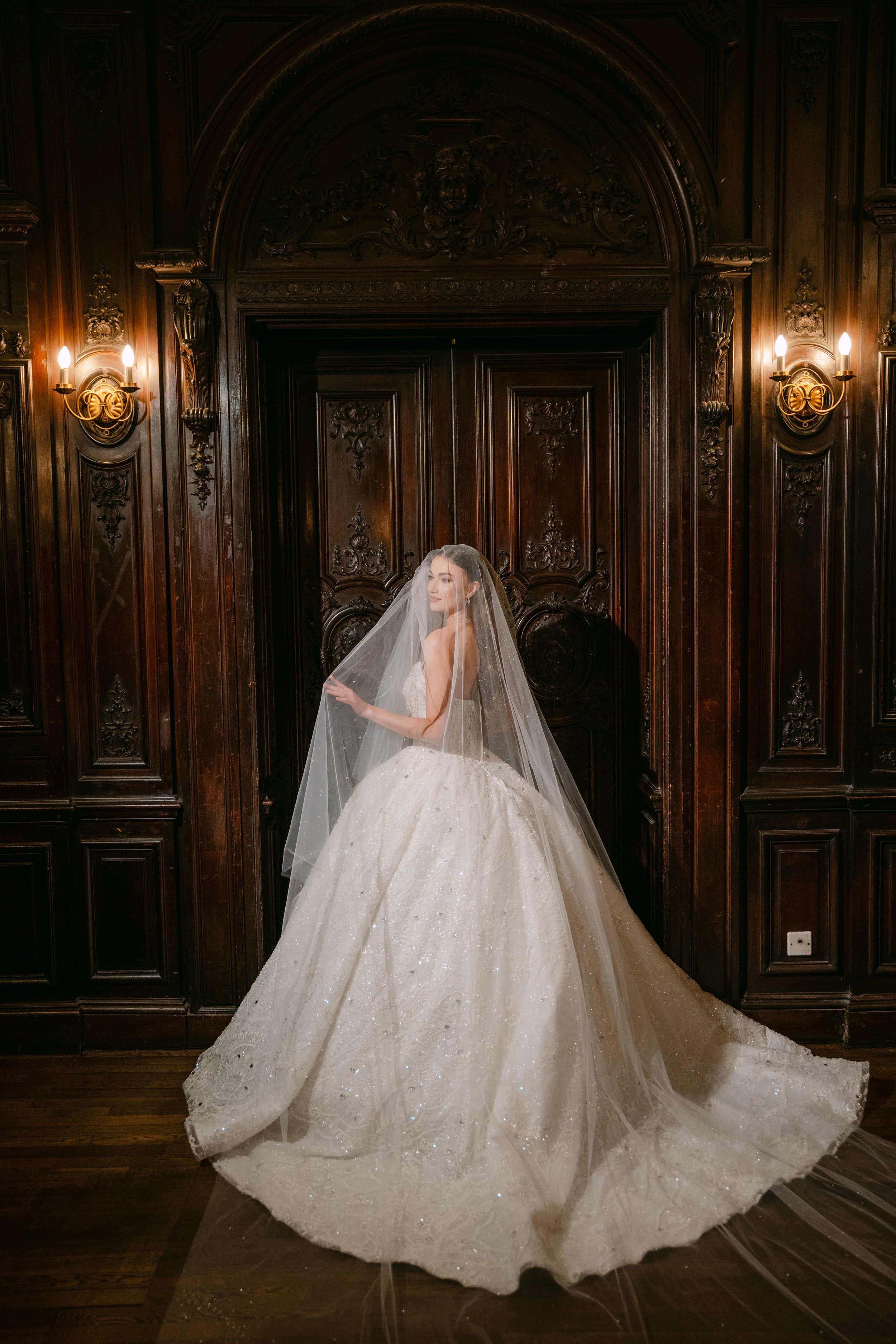 A woman in a white wedding gown and veil stands in a dark, ornate room with wooden paneling and wall sconces.