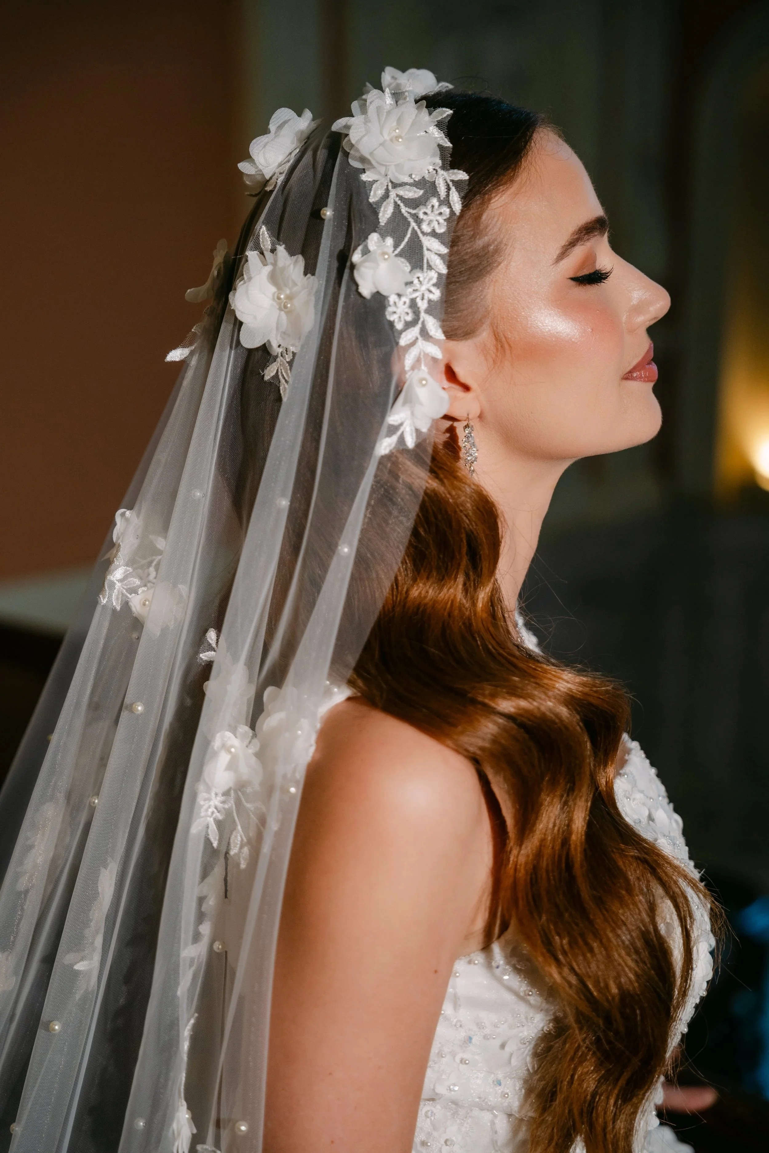 Bridal hair and makeup in a wedding dress with lace and pearl details, wearing a floral embroidered veil and dangling earrings, with her eyes closed and smiling subtly.