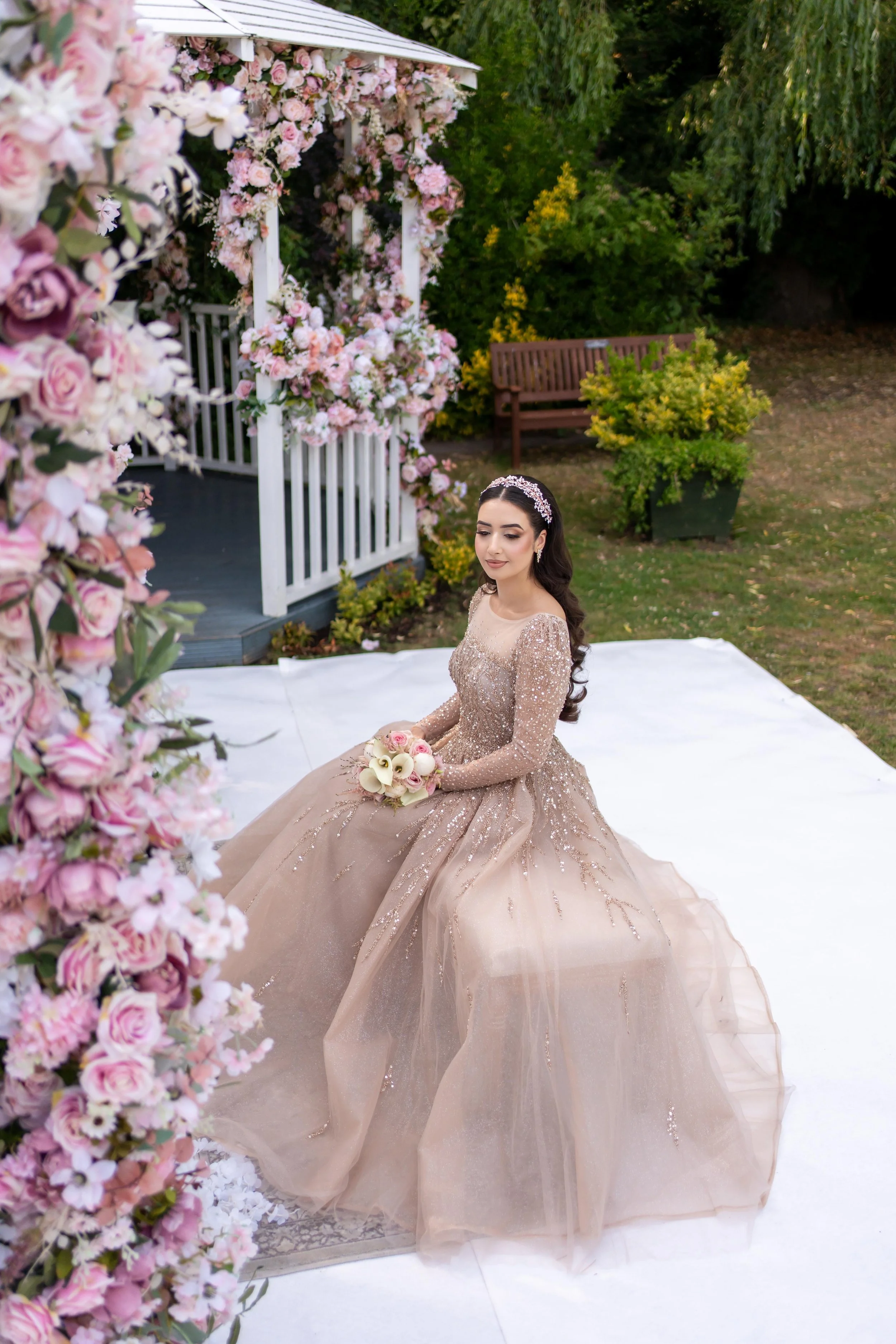 A woman in a sparkling beige gown sitting outdoors on a white cloth, holding a bouquet of white and pink flowers, with a floral arch, garden, and trees in the background.