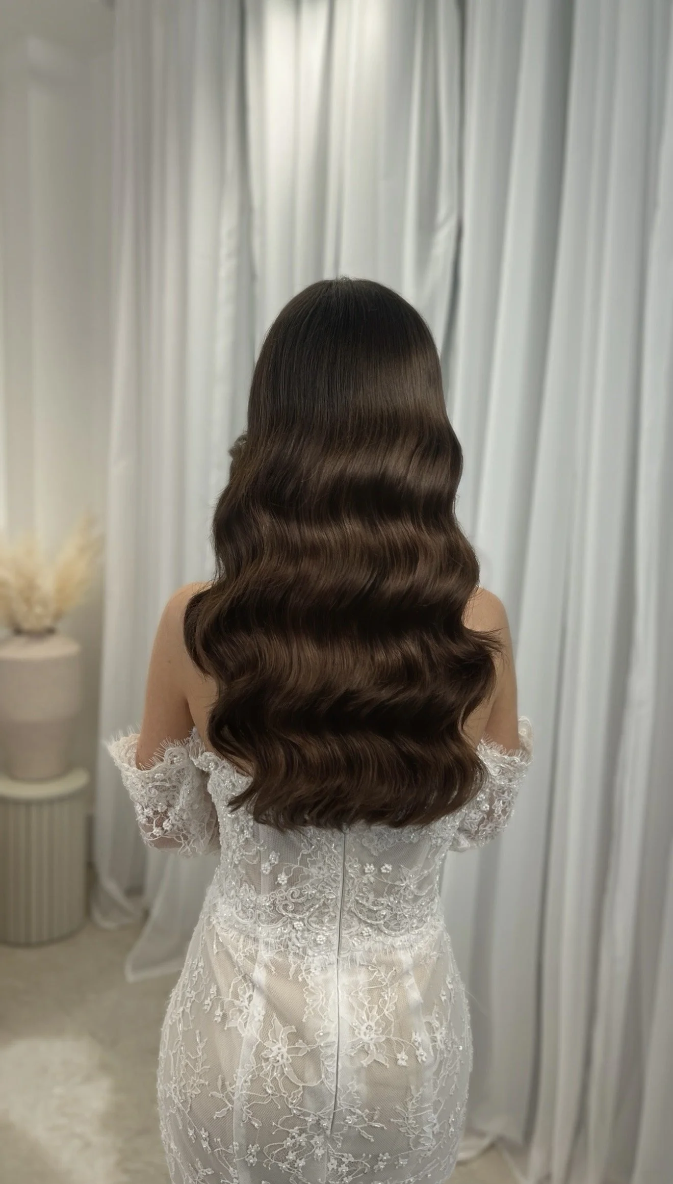 Back view of a woman with long, wavy brown hair wearing a white lace off-shoulder dress, standing in front of a light-colored curtain.
