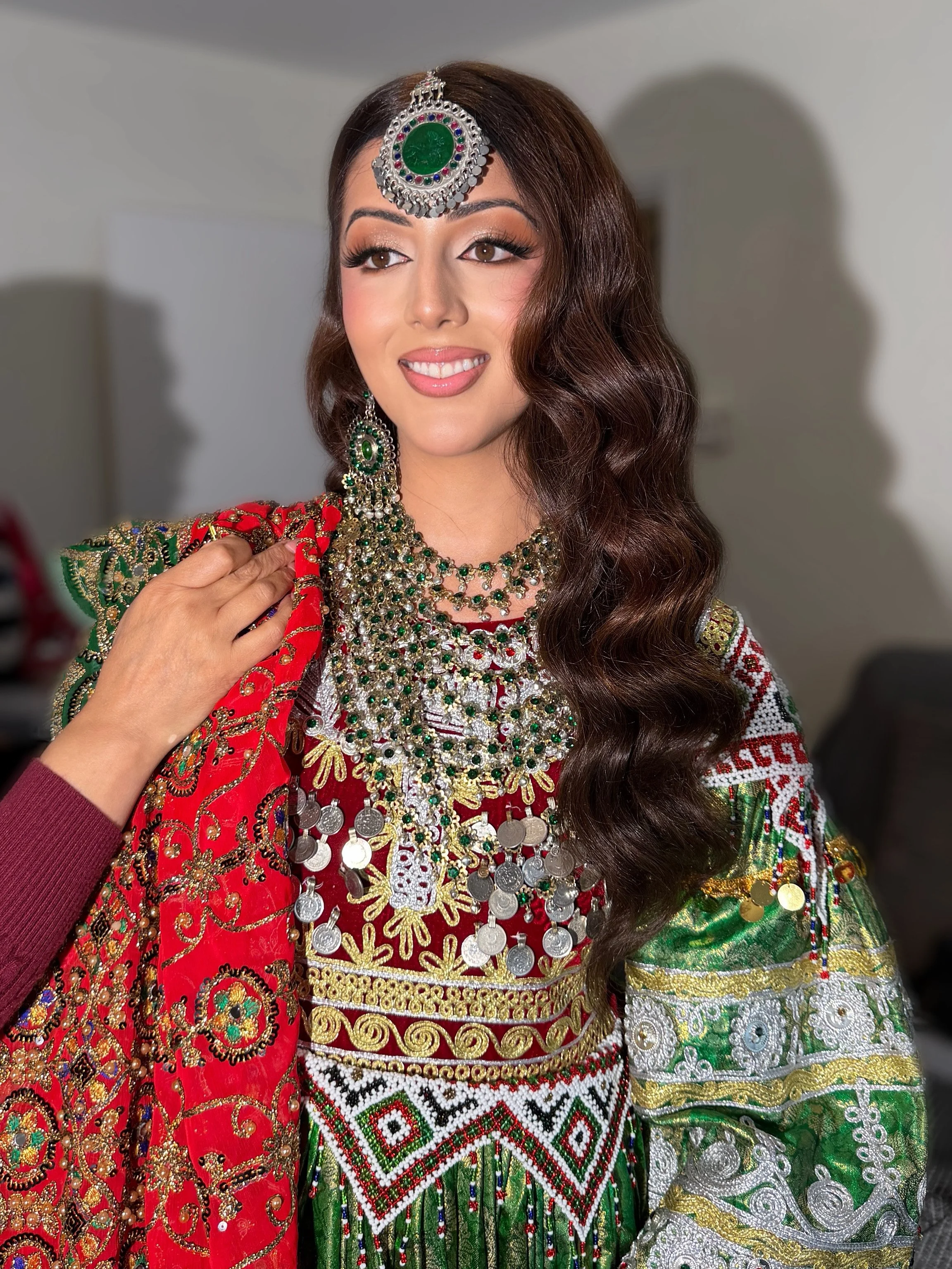 A woman dressed in traditional colorful attire with elaborate jewelry, including a headpiece, earrings, and layered necklaces. She has long wavy hair and is smiling while looking to the side.