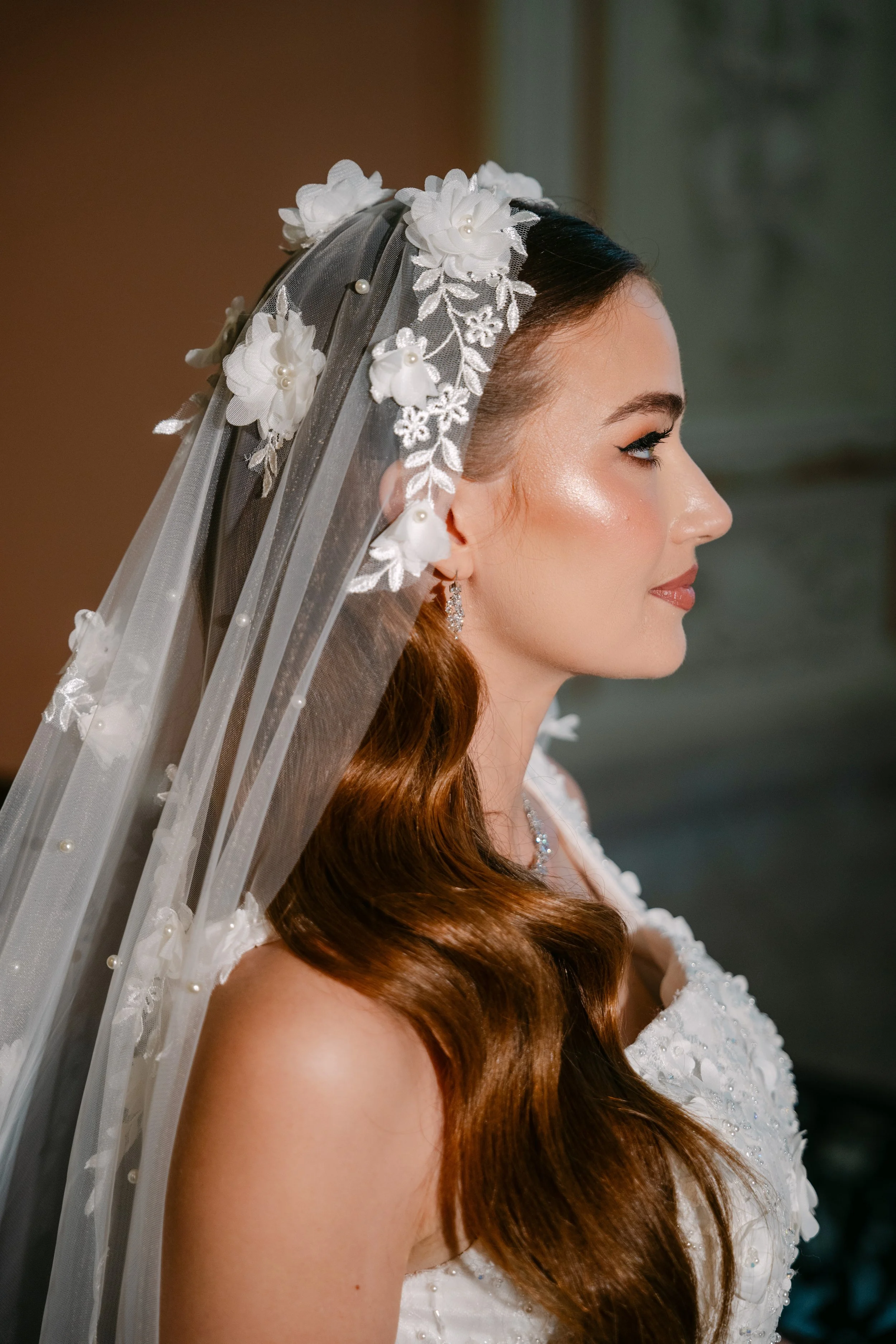 Profile view of a woman with long, wavy, auburn hair wearing a white lace wedding dress with pearls and a bridal veil decorated with fabric flowers and pearls.