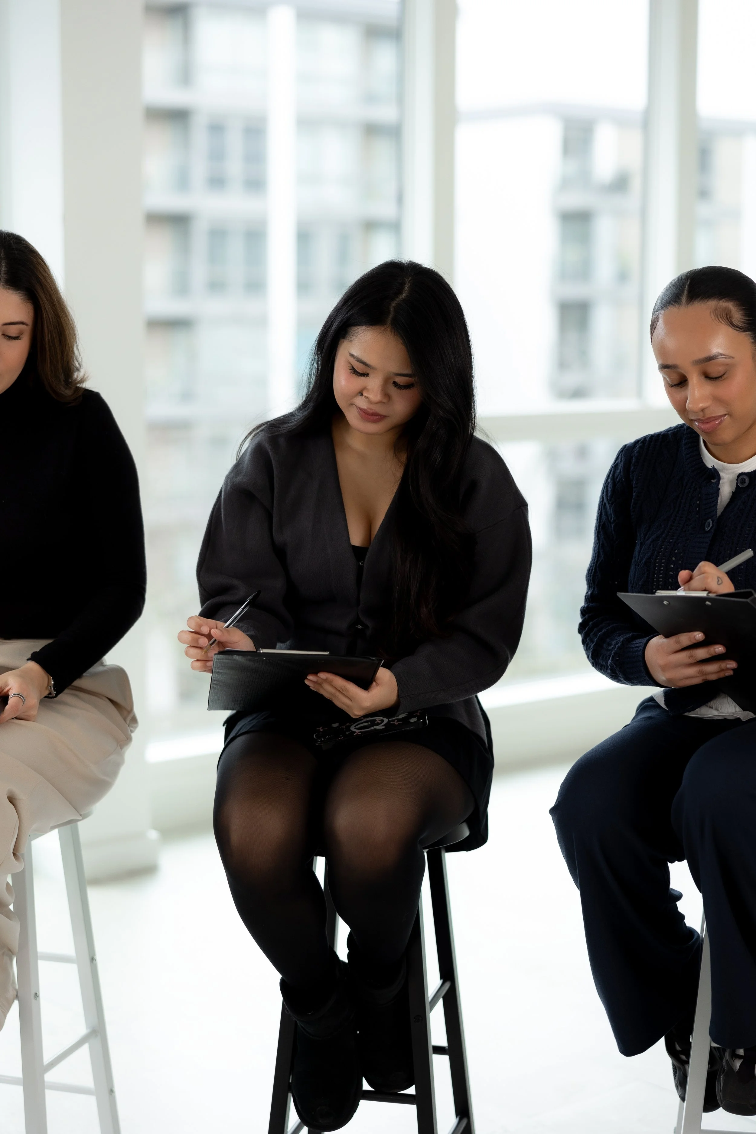 Three women sitting in a row, one holding a clipboard and pen, in a bright room with large windows and city buildings in the background.