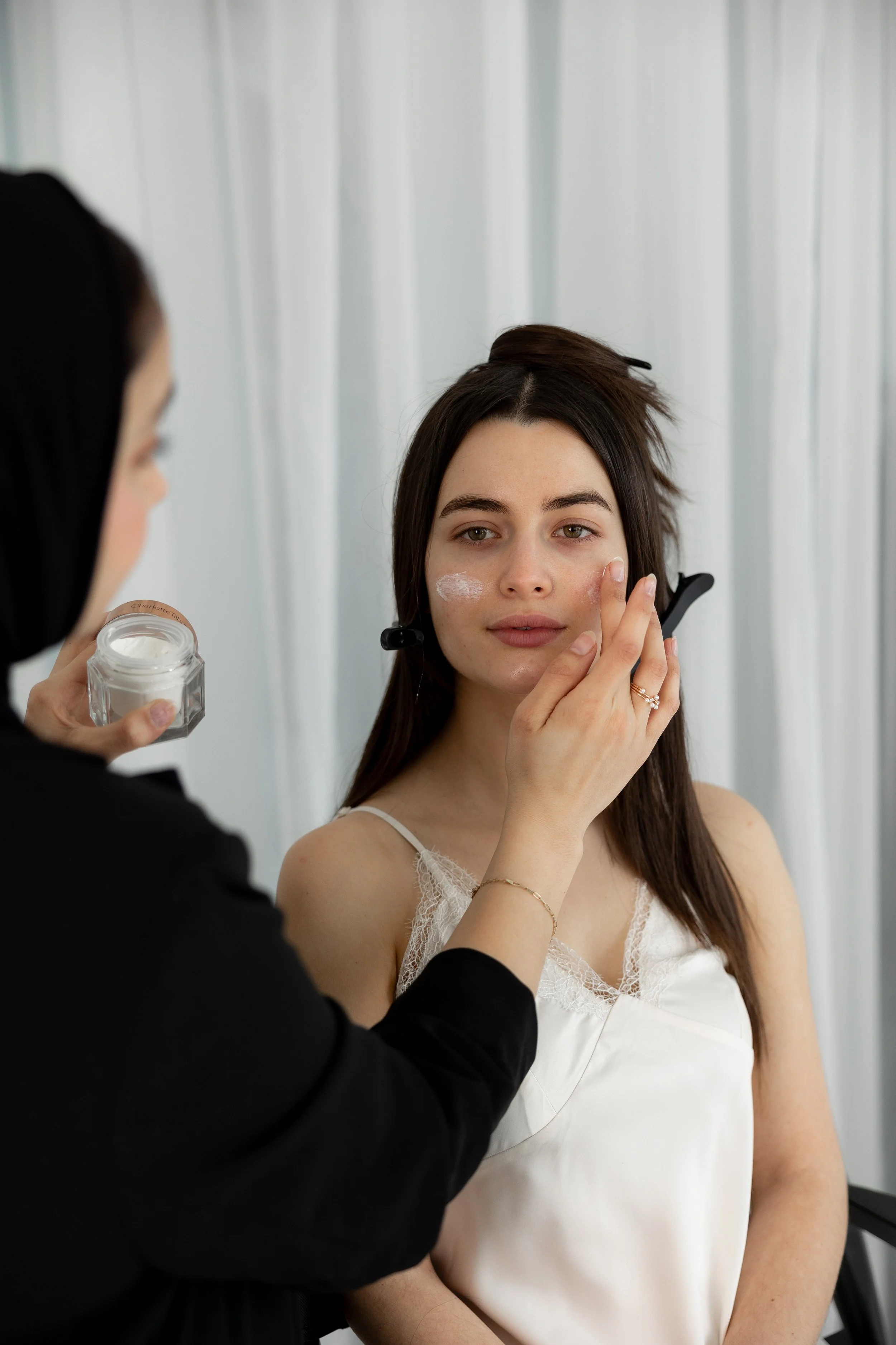 A woman with long dark hair, wearing a white lace top, is getting makeup applied around her face by a person holding a small jar of white cream. The setting appears to be a makeup studio with a light-colored curtain backdrop.