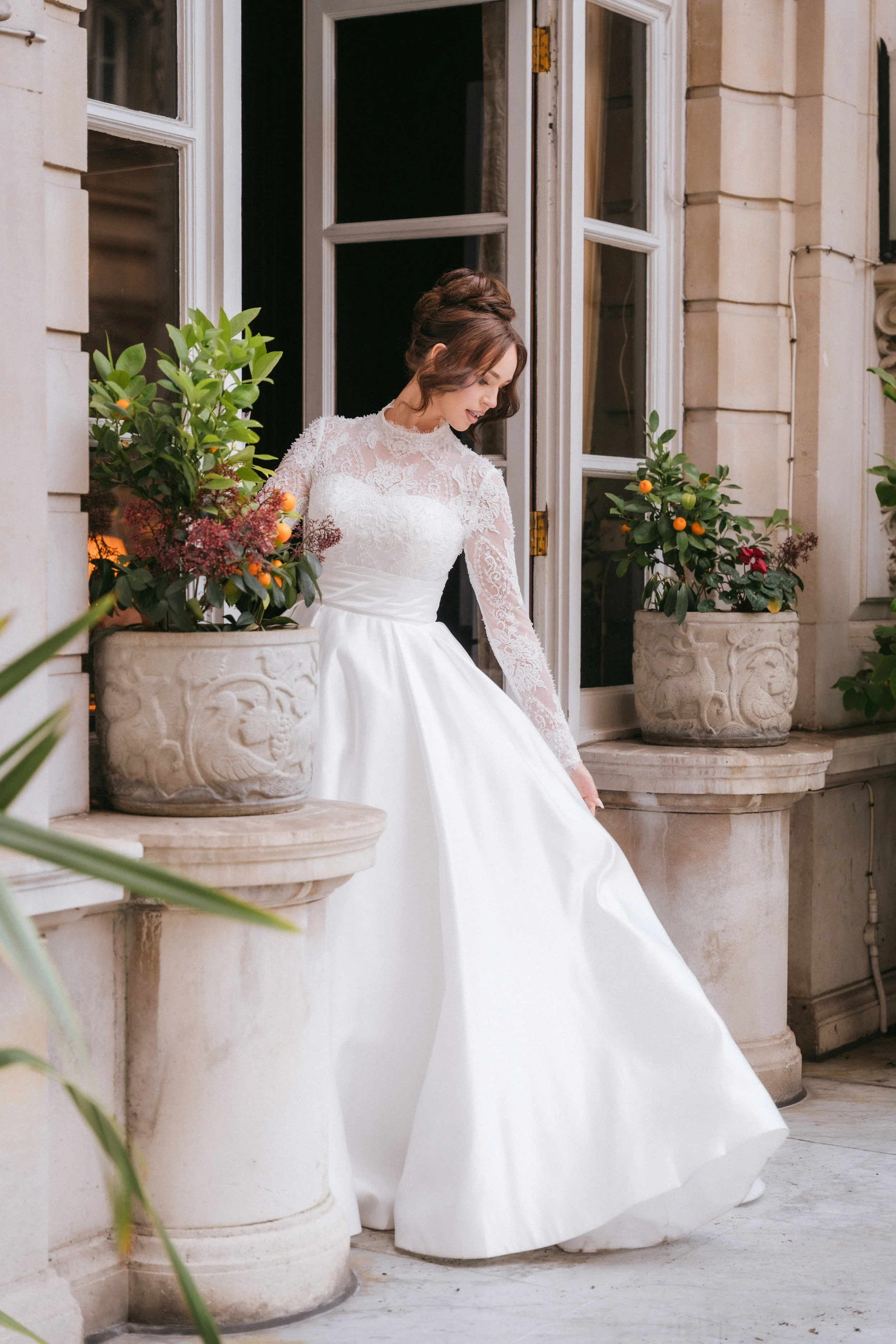 A woman dressed in a white wedding gown with lace sleeves and high bun hairstyle, sitting on a stone ledge between two large planters with green plants and small orange fruits, near a building with large windows and stone exterior.