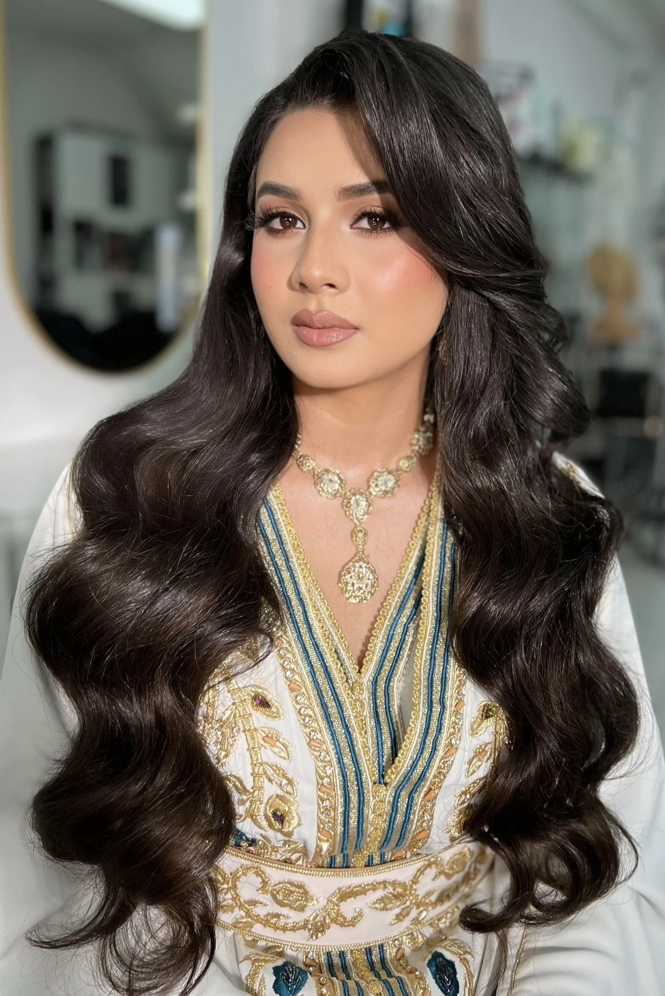 Bridal Hollywood waves By Crown, wearing a white and gold embroidered traditional dress and a gold necklace, standing in a well-lit indoor space.