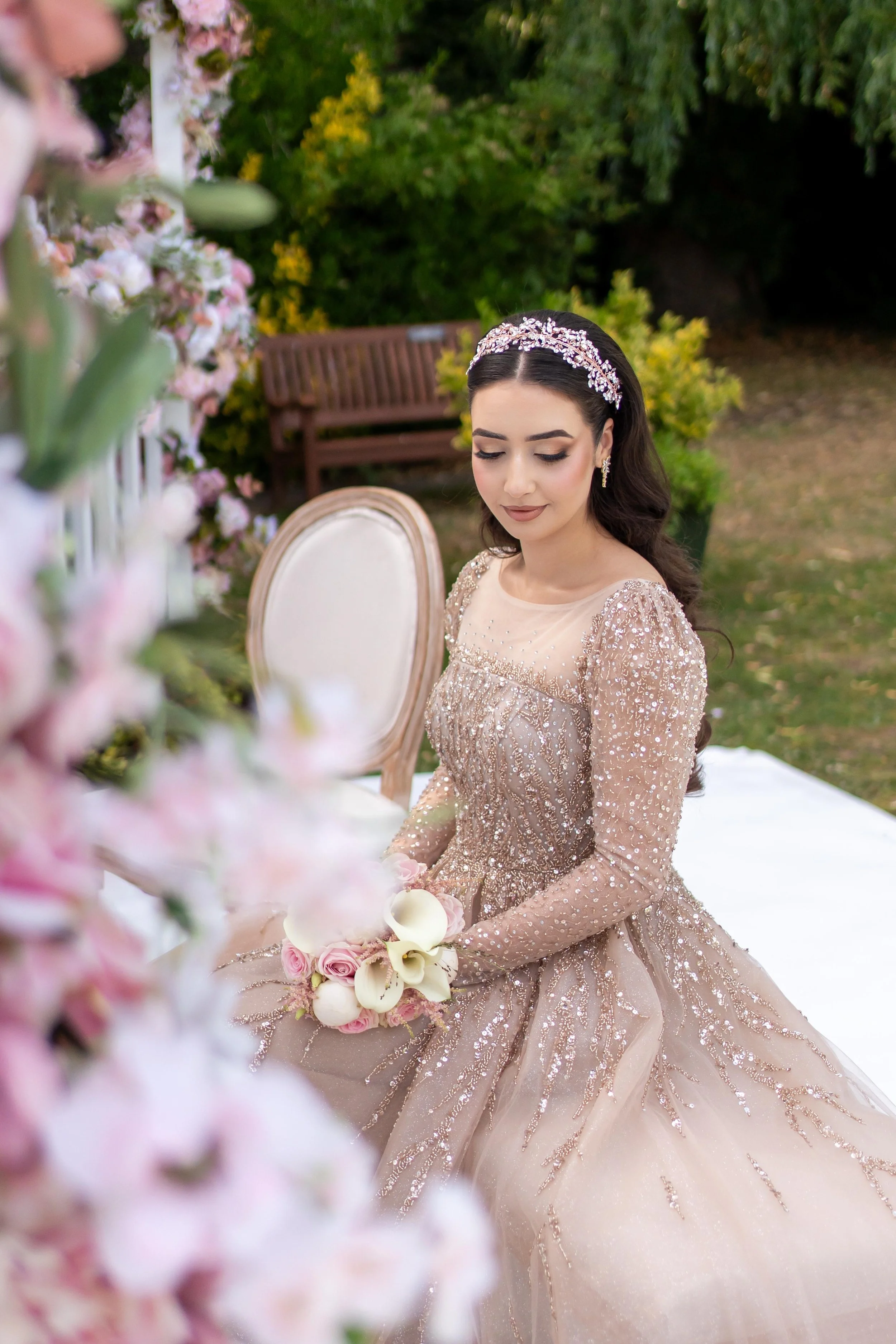A woman in a fancy, embellished gown with long sleeves and a full skirt, sitting outdoors at a table decorated with pink flowers and holding a bouquet of pink roses and white calla lilies. She has dark hair styled in waves, adorned with a bejeweled headband, and is wearing earrings.