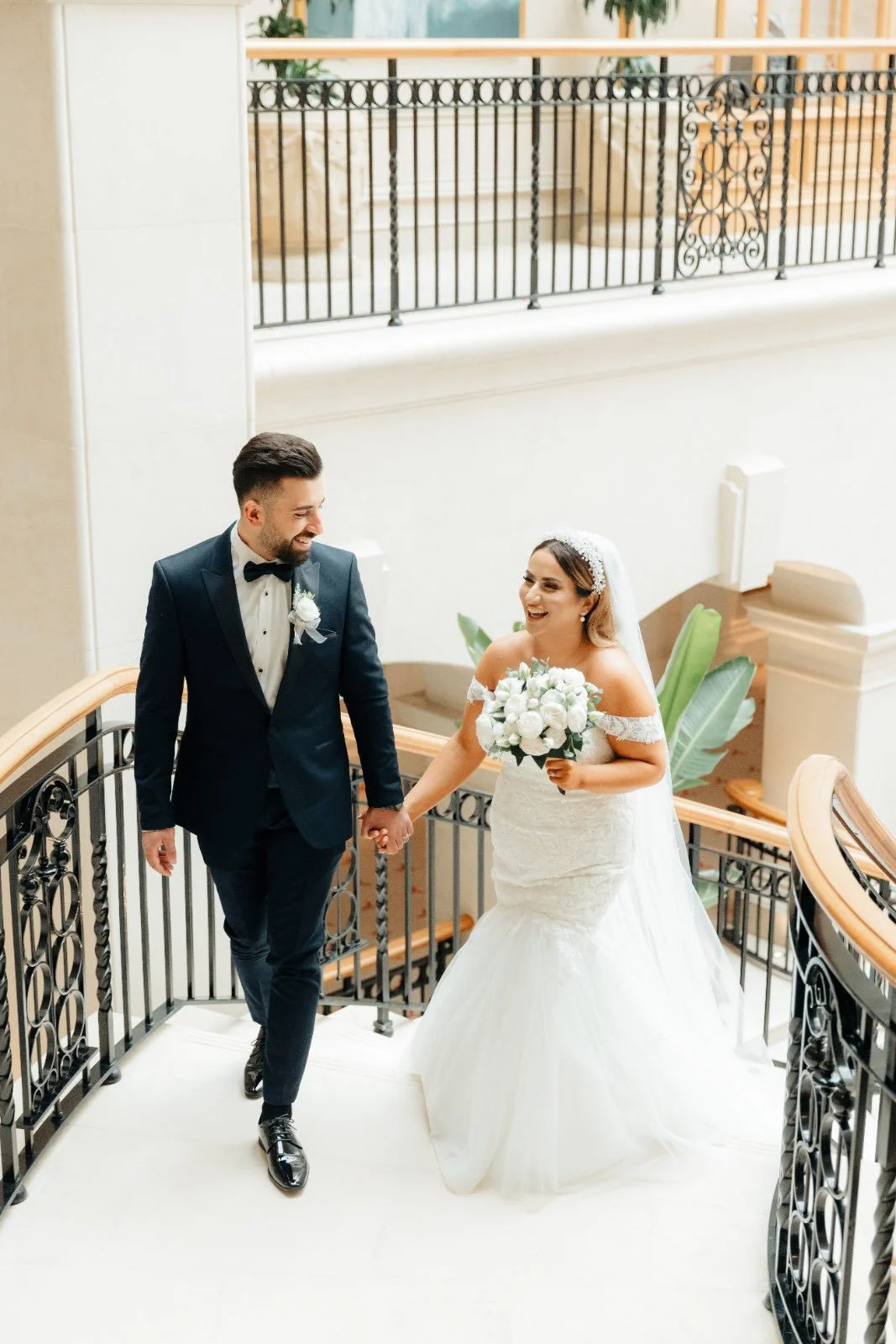 A newlywed couple holding hands and smiling on a staircase, with the bride in a white wedding gown holding a bouquet of white flowers, and the groom in a black tuxedo with a white boutonniere.