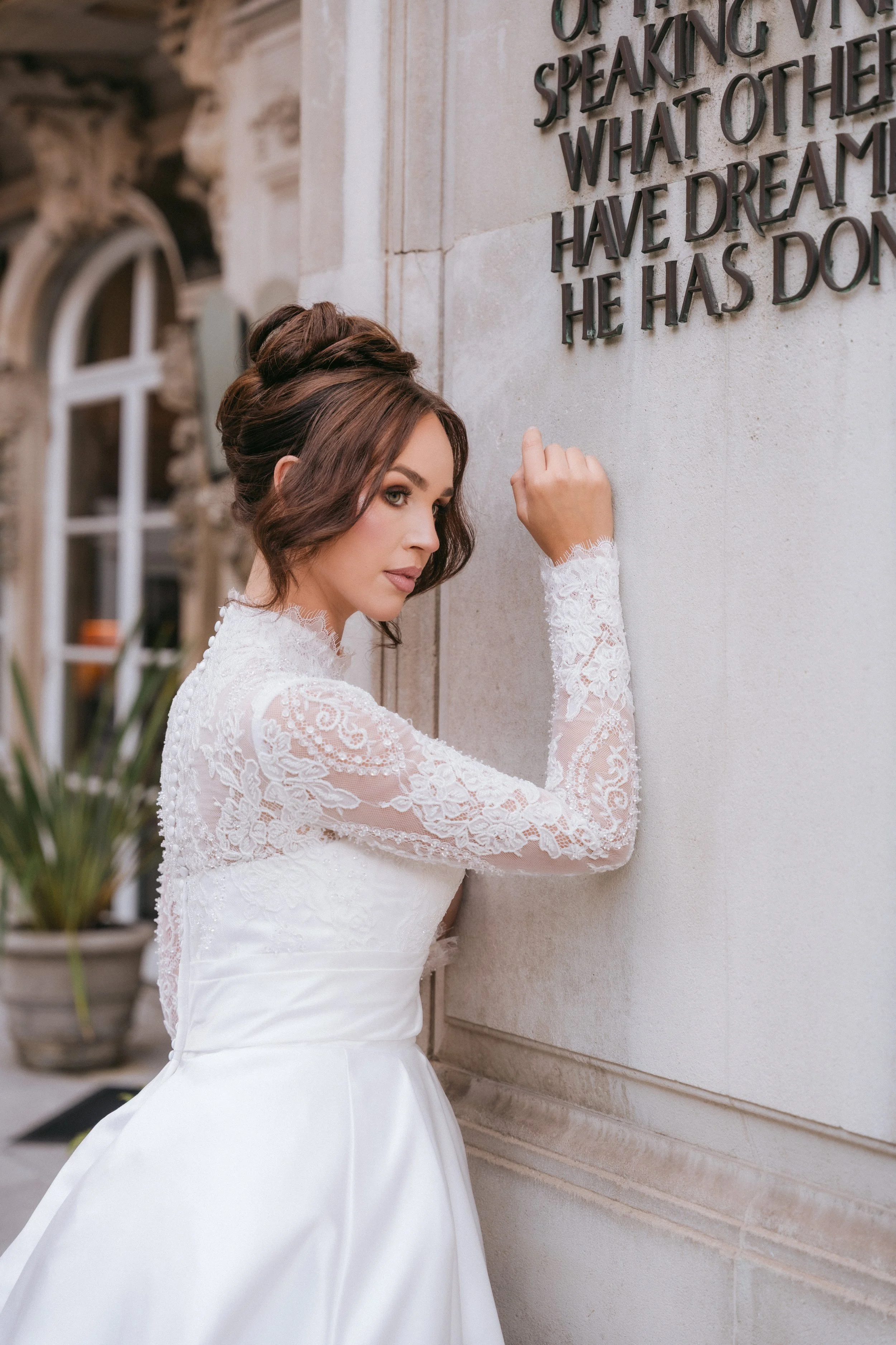 A bride in a white lace wedding dress with a satin skirt, standing by a stone wall with a metal plaque containing engraved text. She has shoulder-length brown hair styled in soft waves, looking slightly to the side with a serious expression.