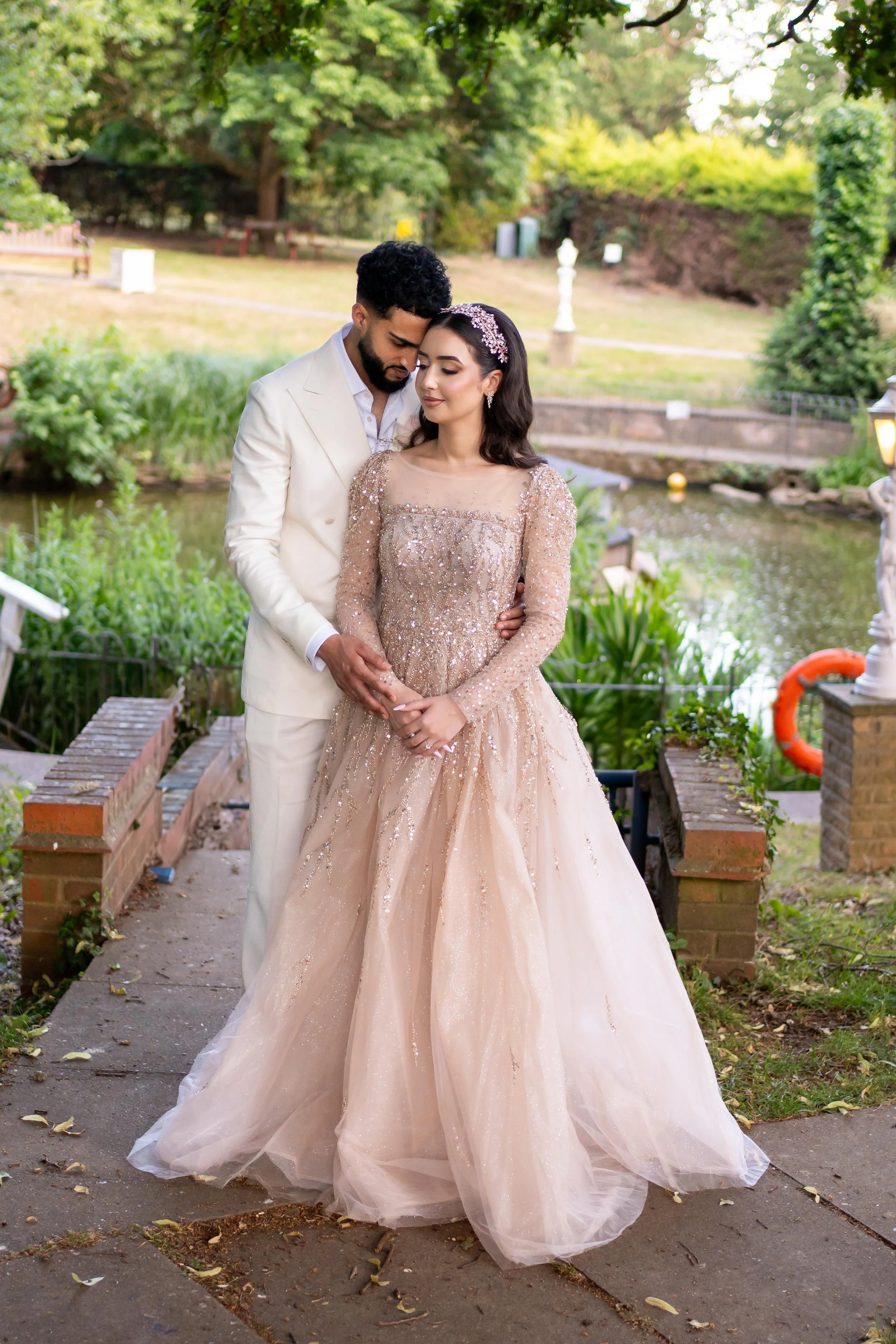 A man and woman dressed in formal attire standing close together outdoors in a garden setting, with the man gently holding the woman's hands and leaning his head towards her, both with peaceful expressions.