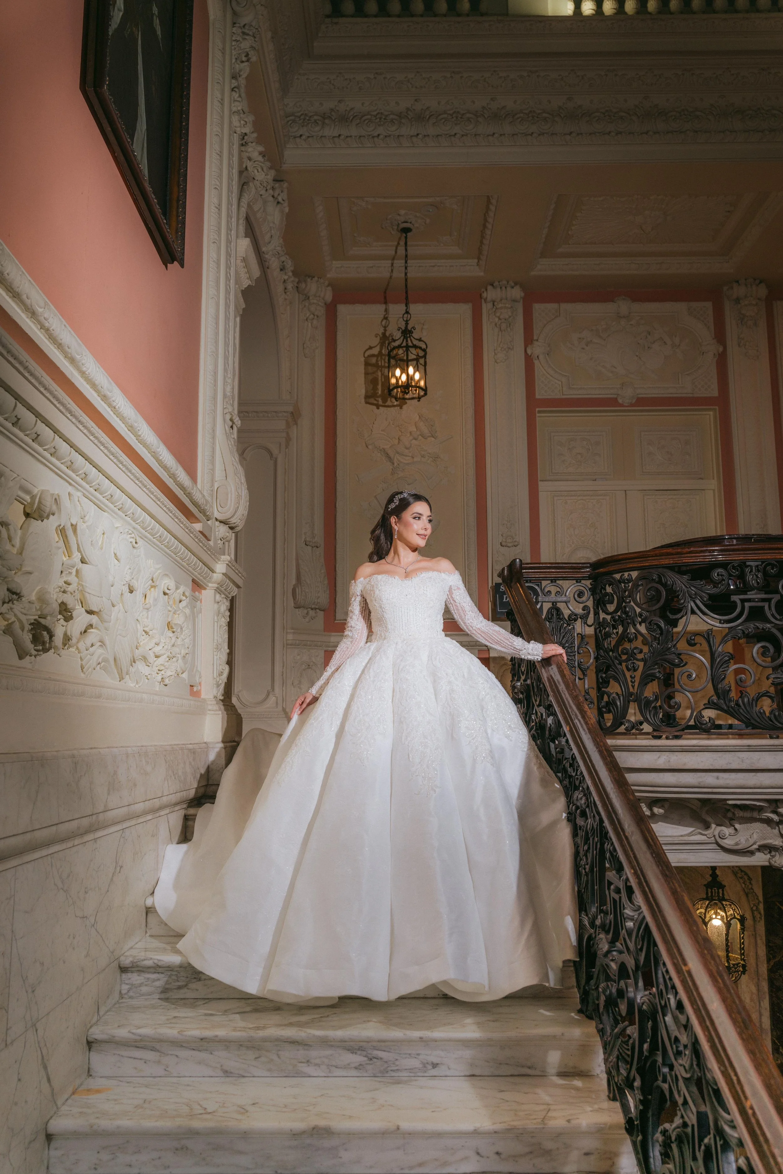 A woman in a white wedding gown standing on a marble staircase in an ornate, historic building with intricate molding and artwork on the walls.