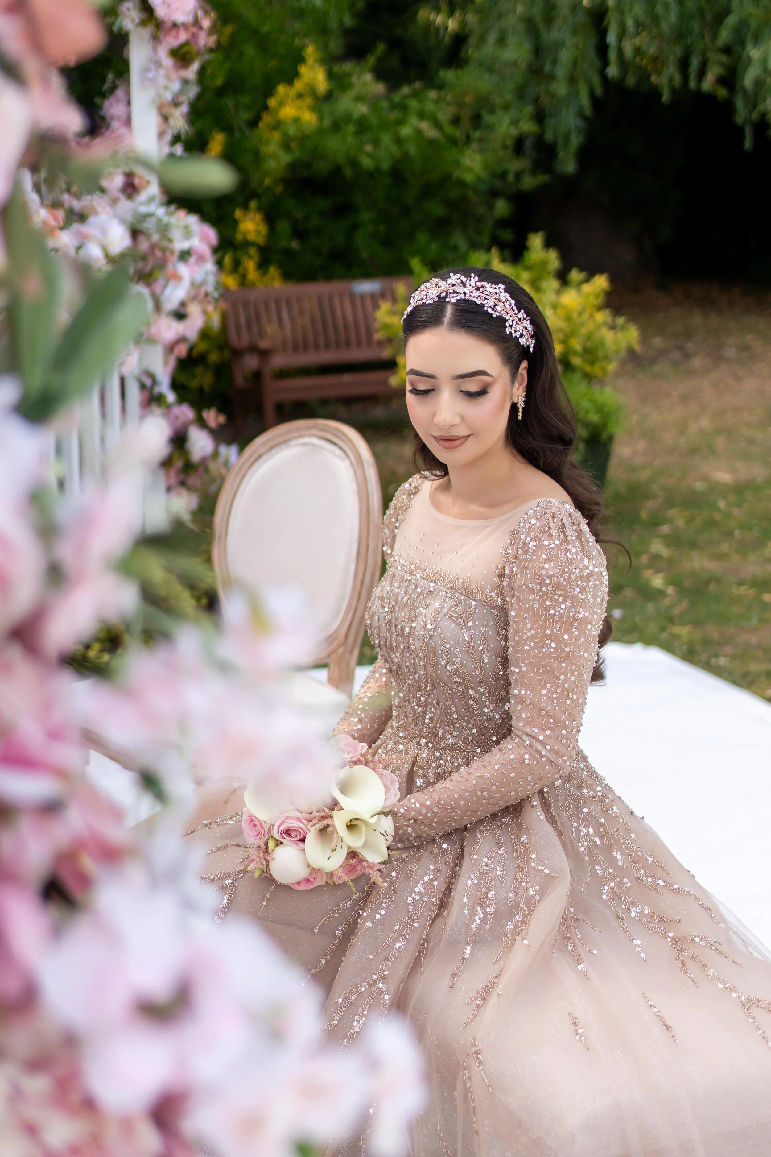 Young woman in a sparkling beige gown with a tiara, sitting outdoors at a wedding or formal event, with pink and white flowers in the foreground and green foliage in the background.