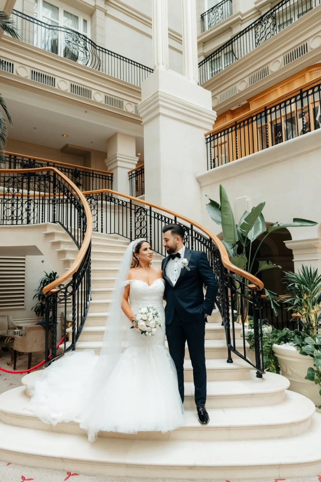 A bride and groom standing together on a staircase inside a grand hotel, with ornate black railings and large green plants around them.