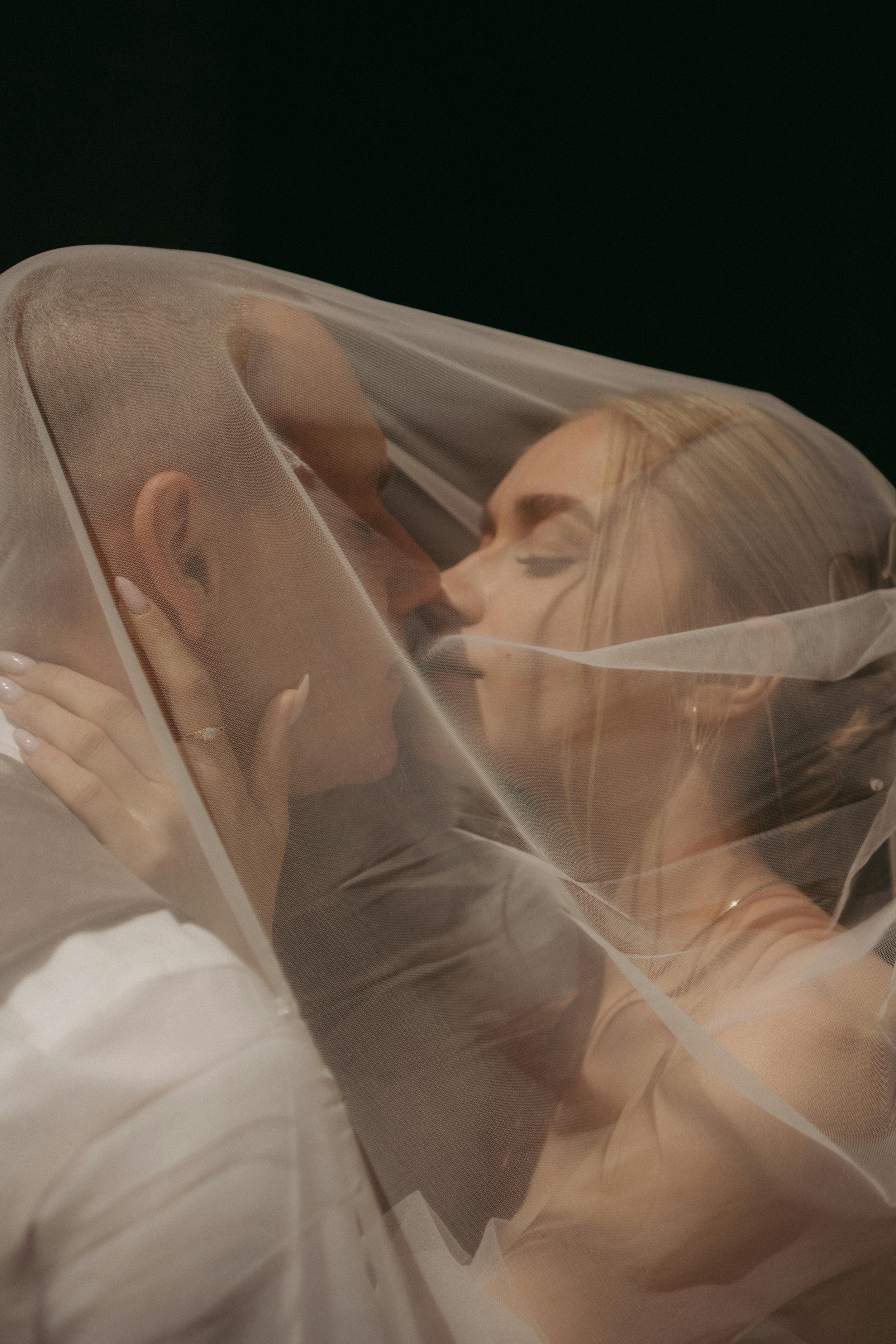 A man and woman covered with a sheer fabric, holding each other closely, with faces nearly touching, in an intimate pose against a dark background.