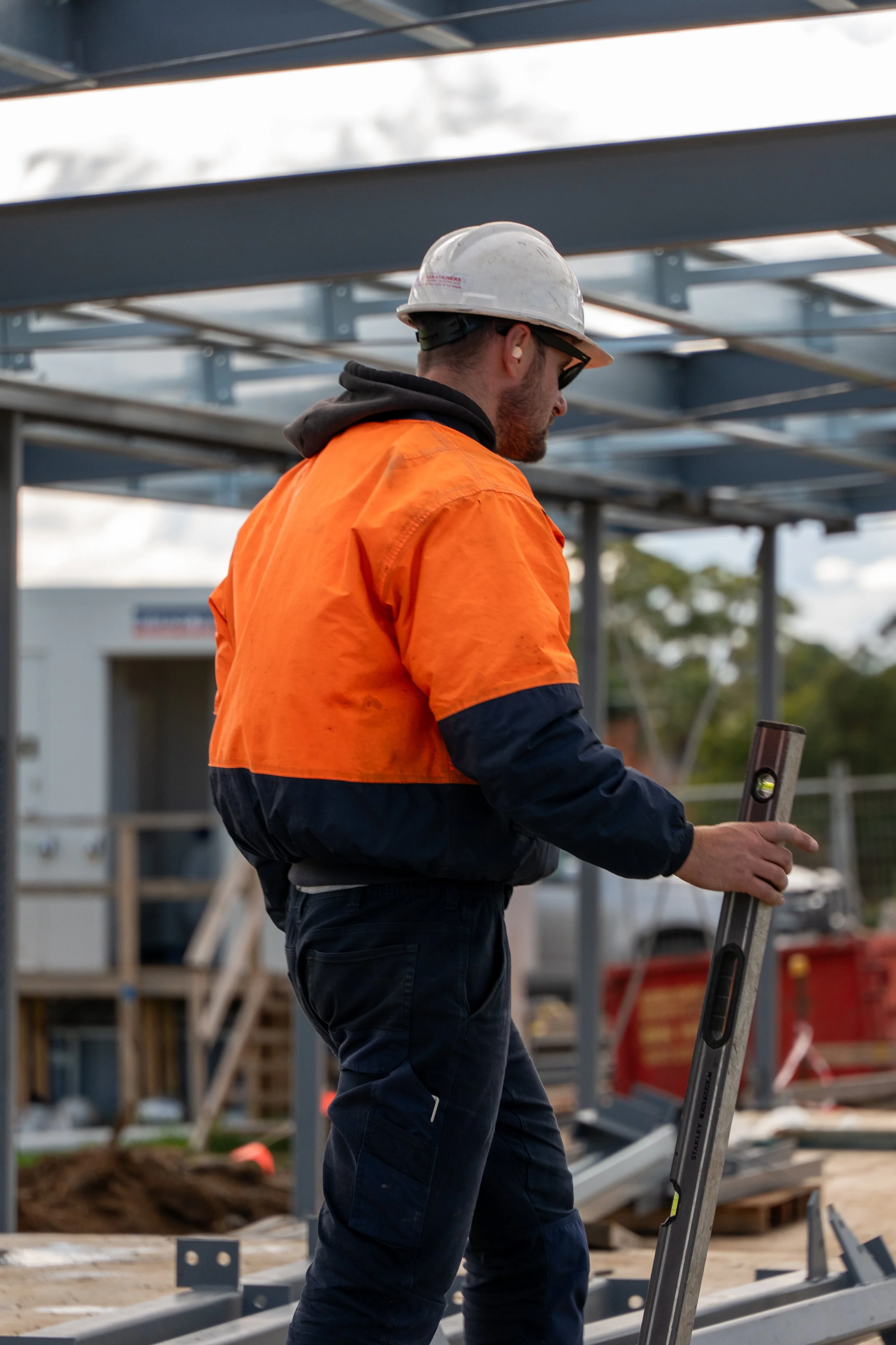 A construction worker wearing an orange safety vest, white hard hat, and sunglasses is using a level tool on a metal structure at a construction site.