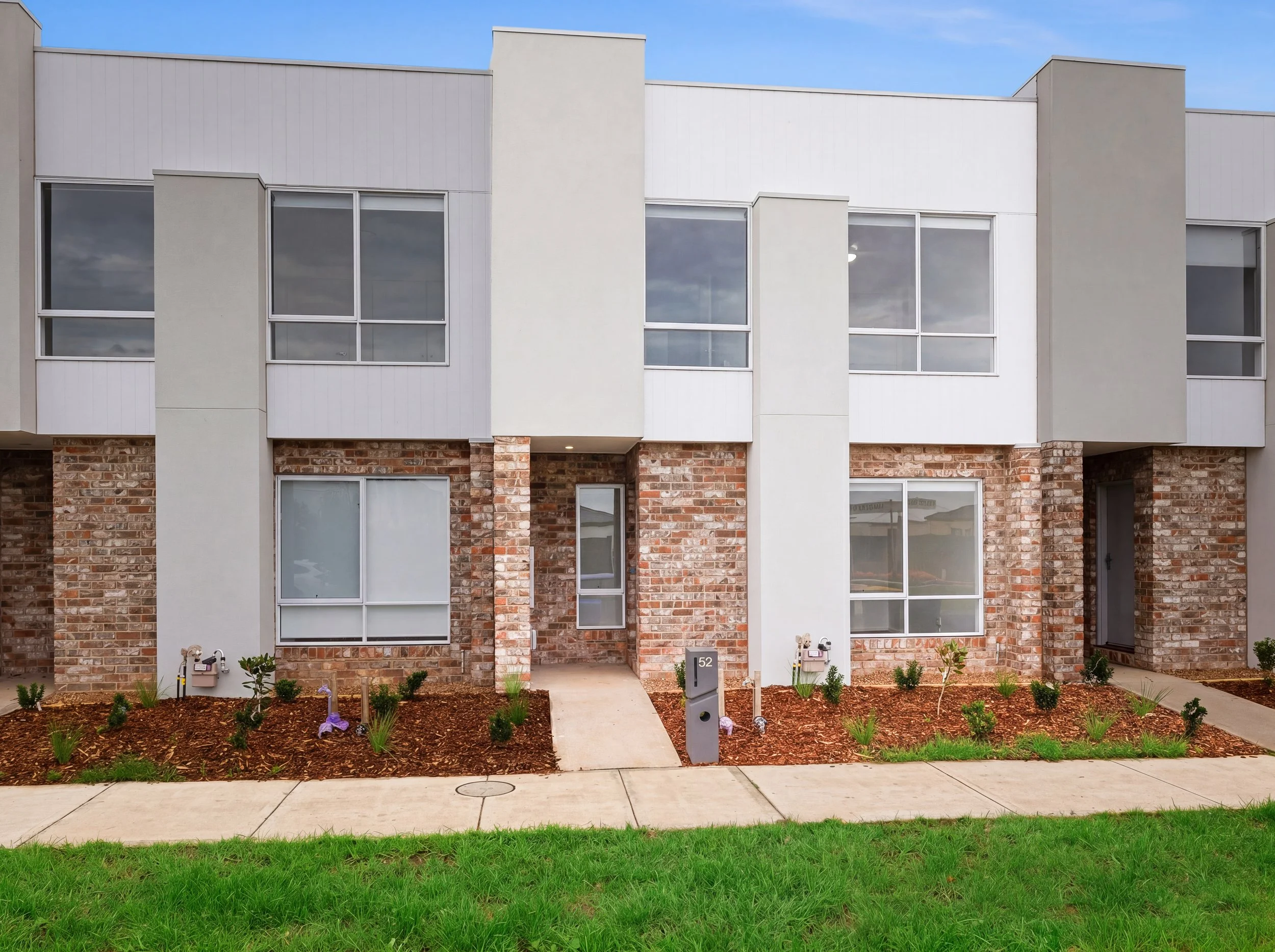 Front view of a modern residential building with a brick and white exterior, large windows, a sidewalk, landscaped garden beds, and a lawn in the foreground.