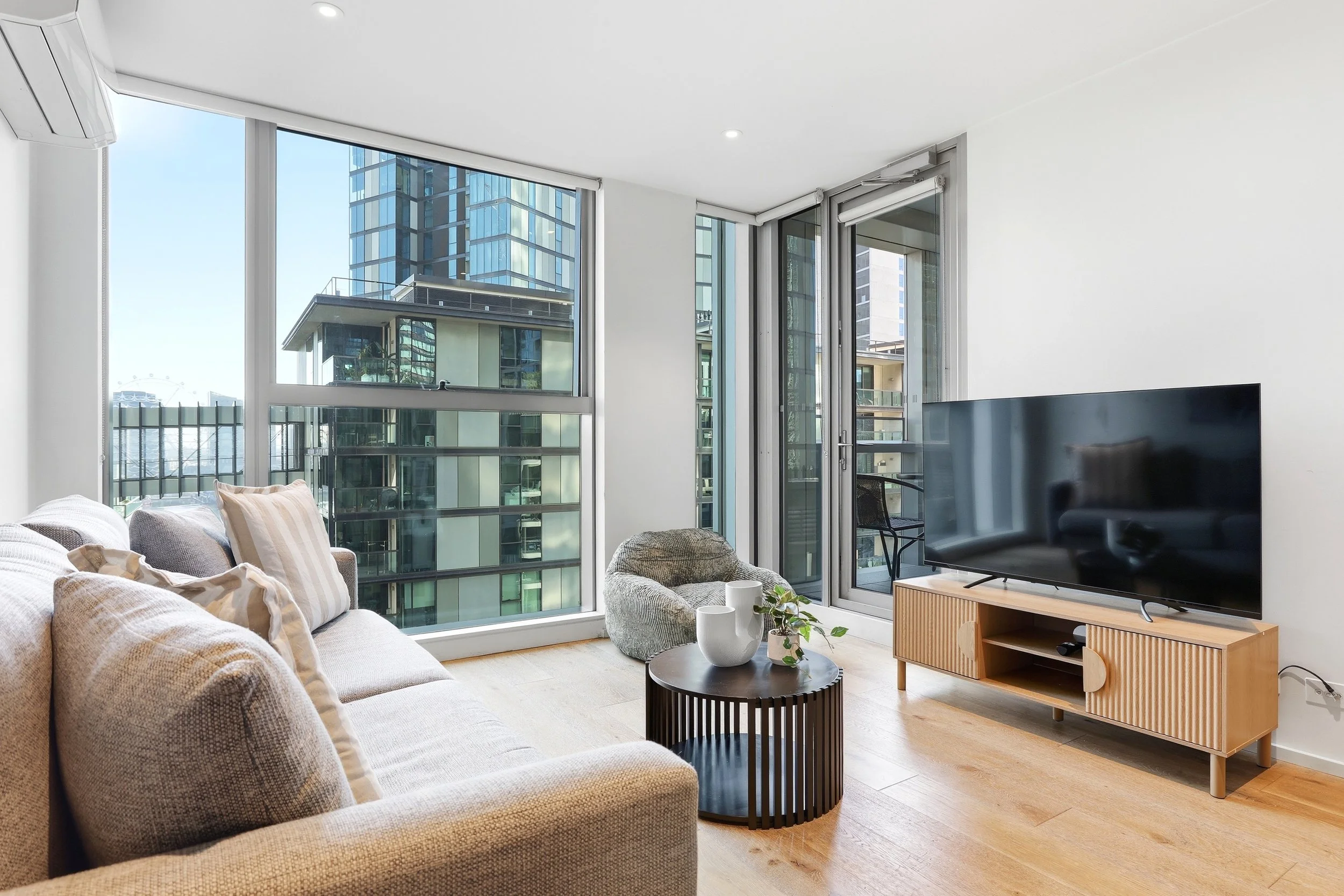 Living room with large floor-to-ceiling windows showing city buildings, a TV on a wooden stand, a beige sofa with pillows, a round black coffee table with plants and cups, a small gray armchair, and wooden flooring.