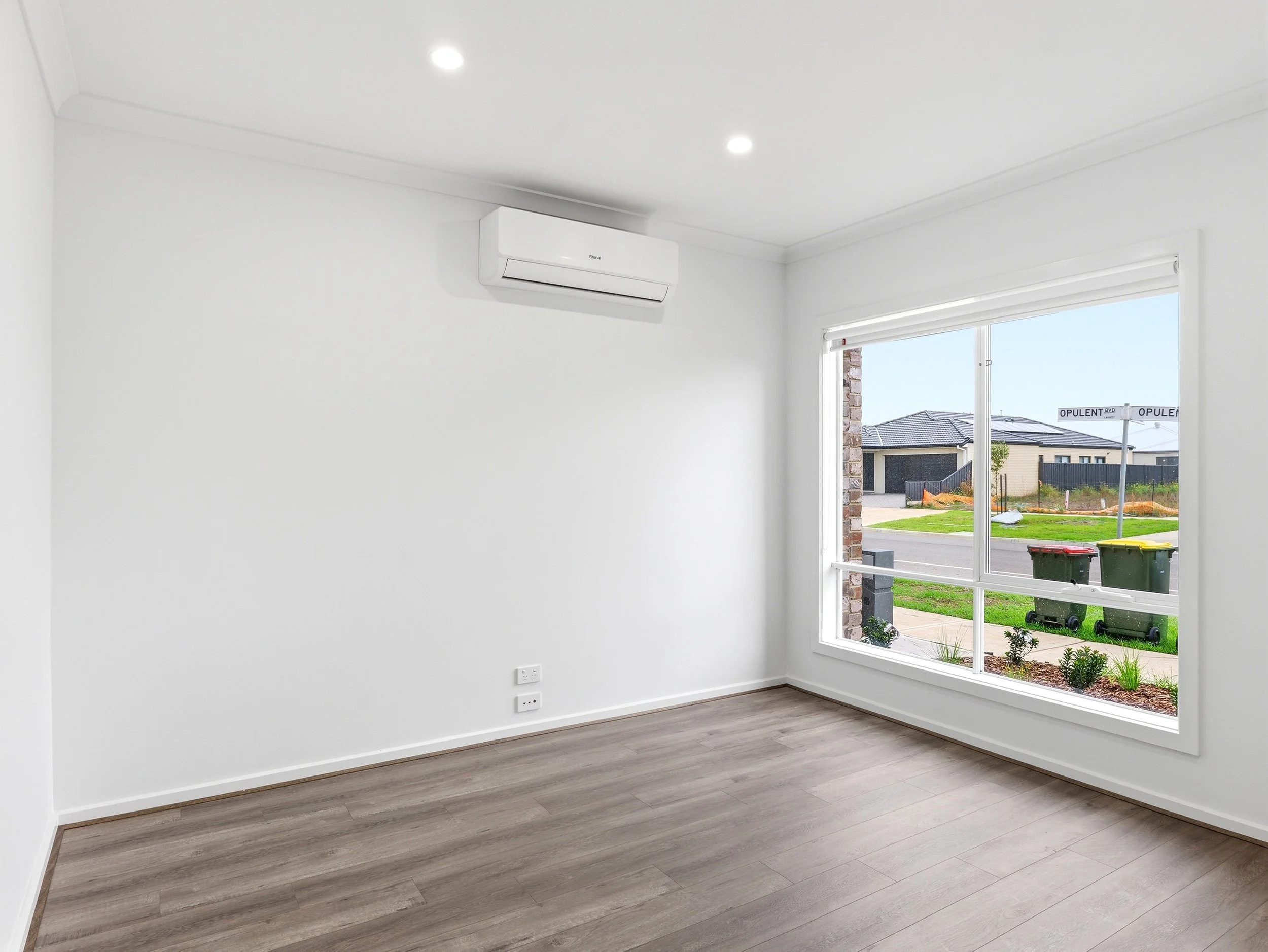 Empty room with white walls, a large window showing a suburban street, and a white wall-mounted air conditioner.