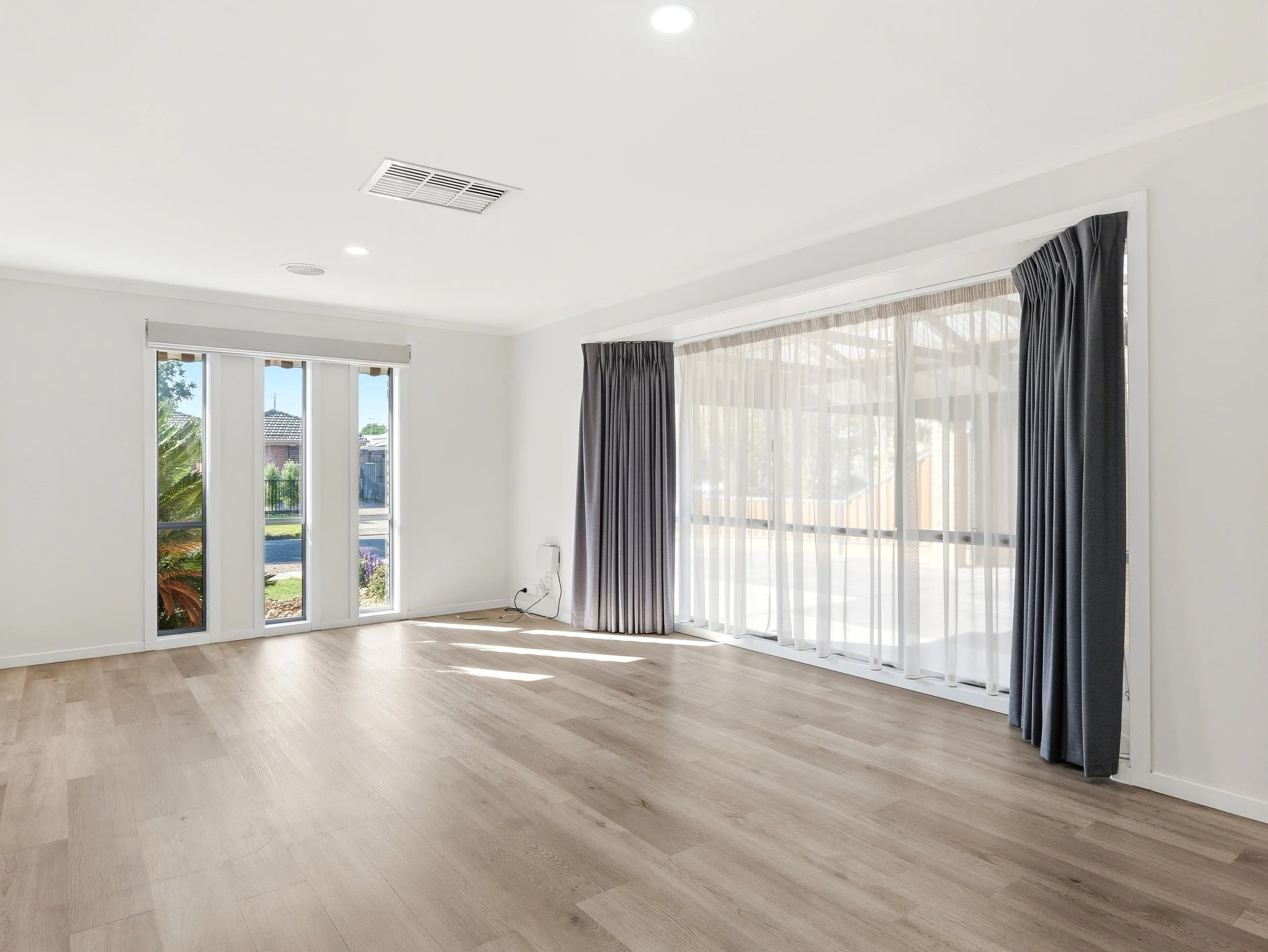 Spacious living room with light wood floors, large windows with sheer and dark curtains, and a white ceiling with recessed lighting and air vent, overlooking a green outdoor area.
