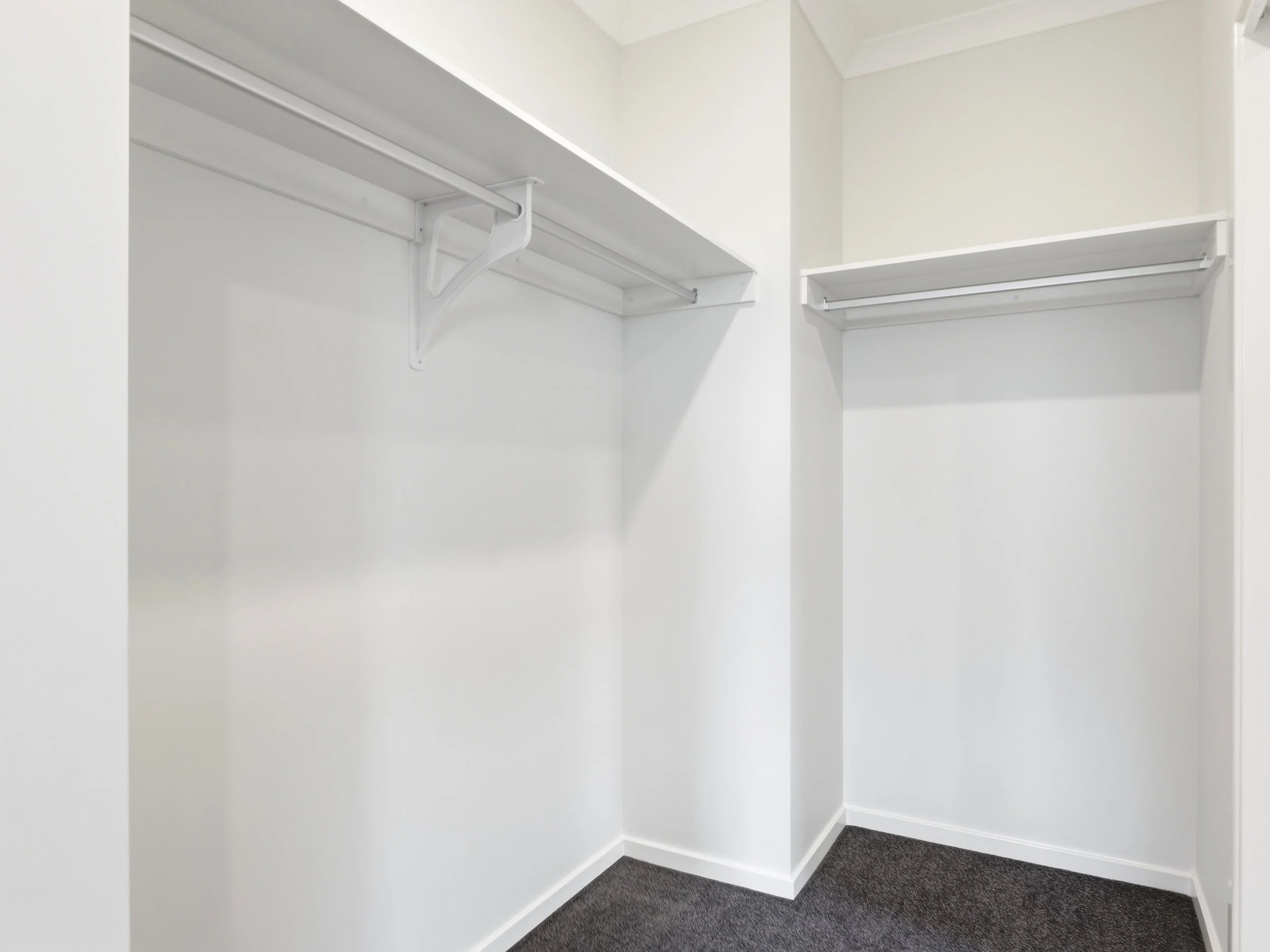 Empty walk-in closet with white walls, hanging rods, and gray carpeted floor.