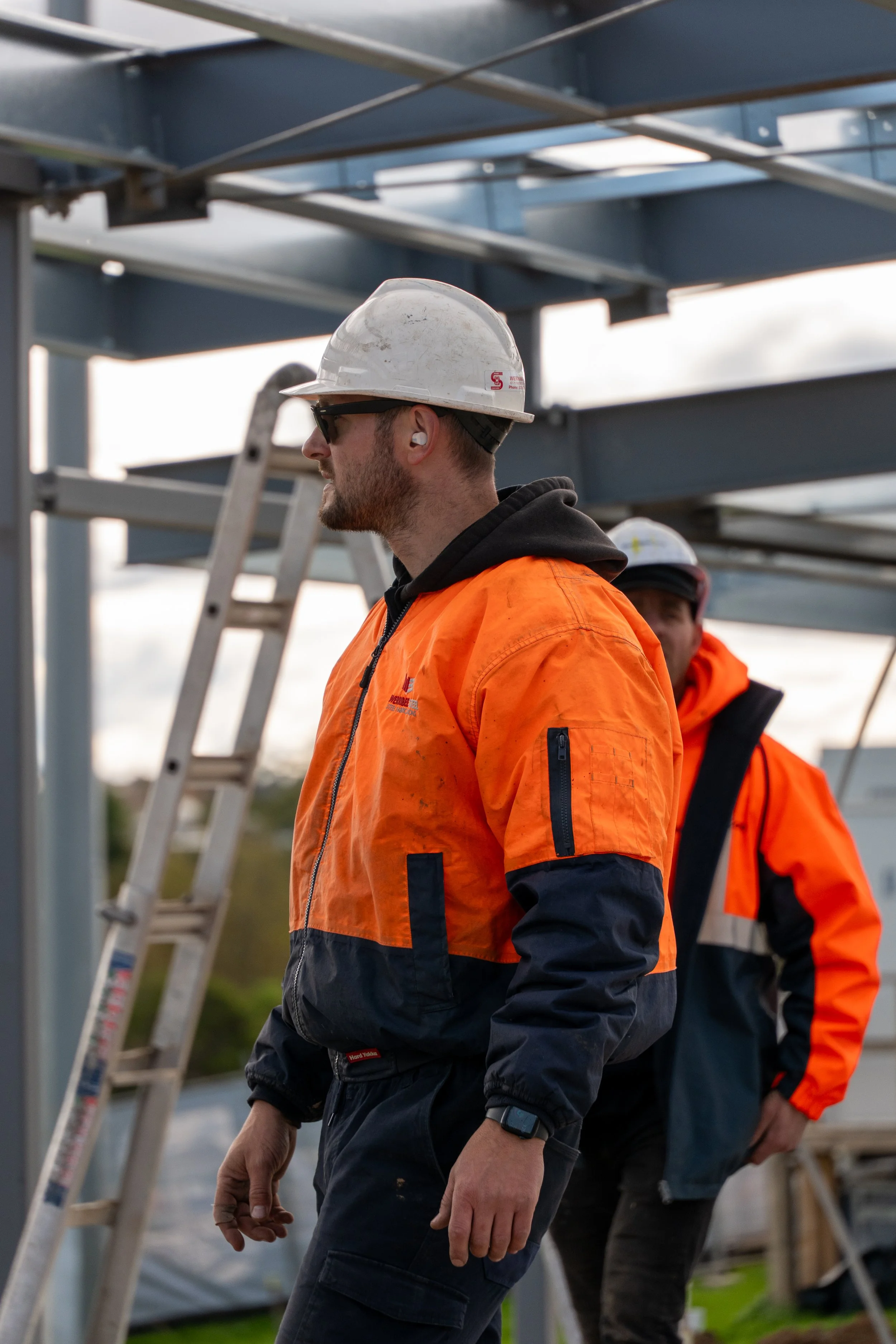 Two construction workers wearing orange safety jackets, white helmets, and protective eyewear are working outdoors near a metal structure. Ladder and construction equipment are visible in the background.