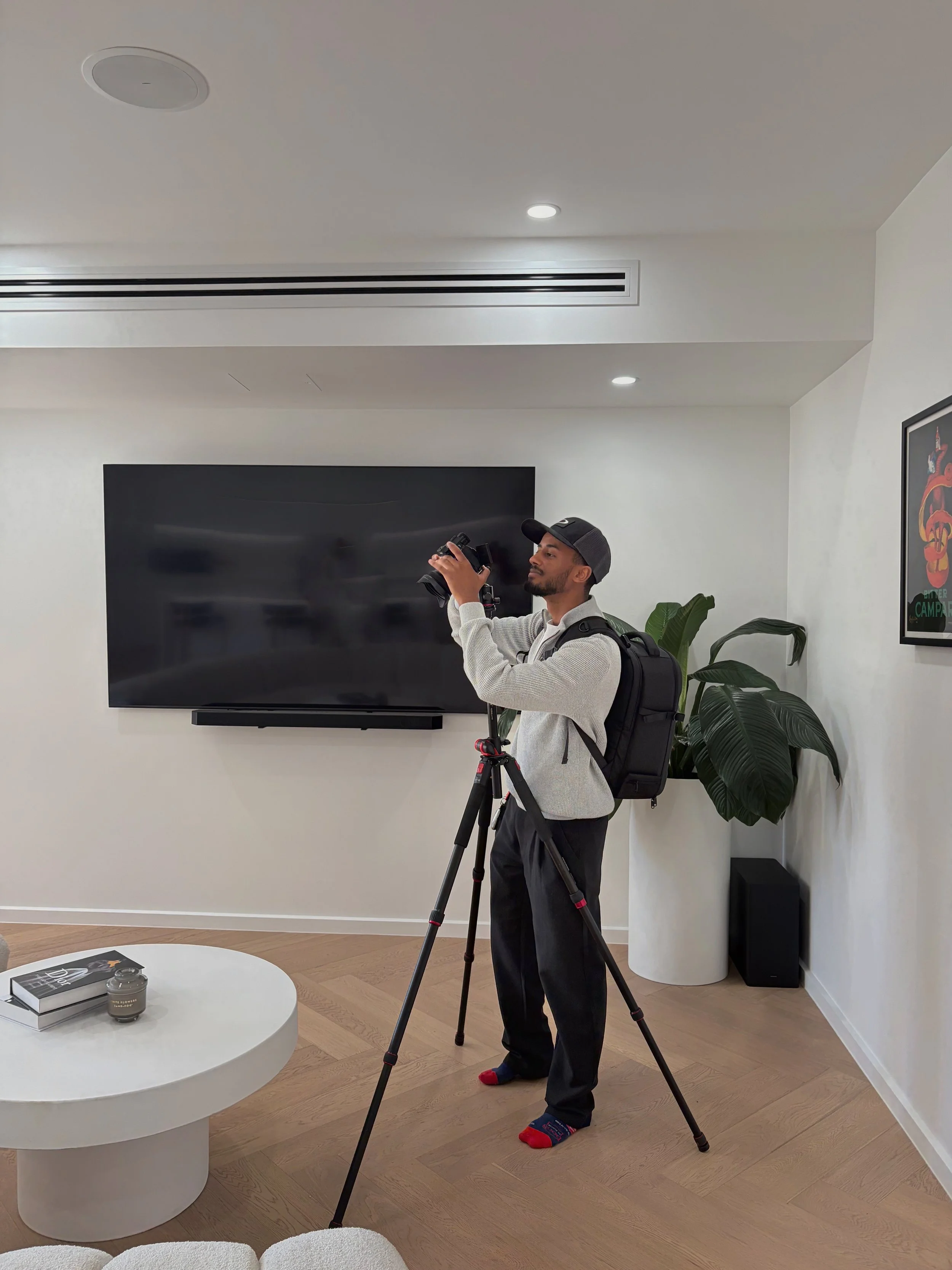 A man standing in a modern living room with a camera on a tripod, wearing a gray sweater, black pants, a baseball cap, and colorful socks, with a backpack on his back. The room has a large flat-screen TV, a potted plant, a framed poster on the wall, and a white circular coffee table with books and a candle.