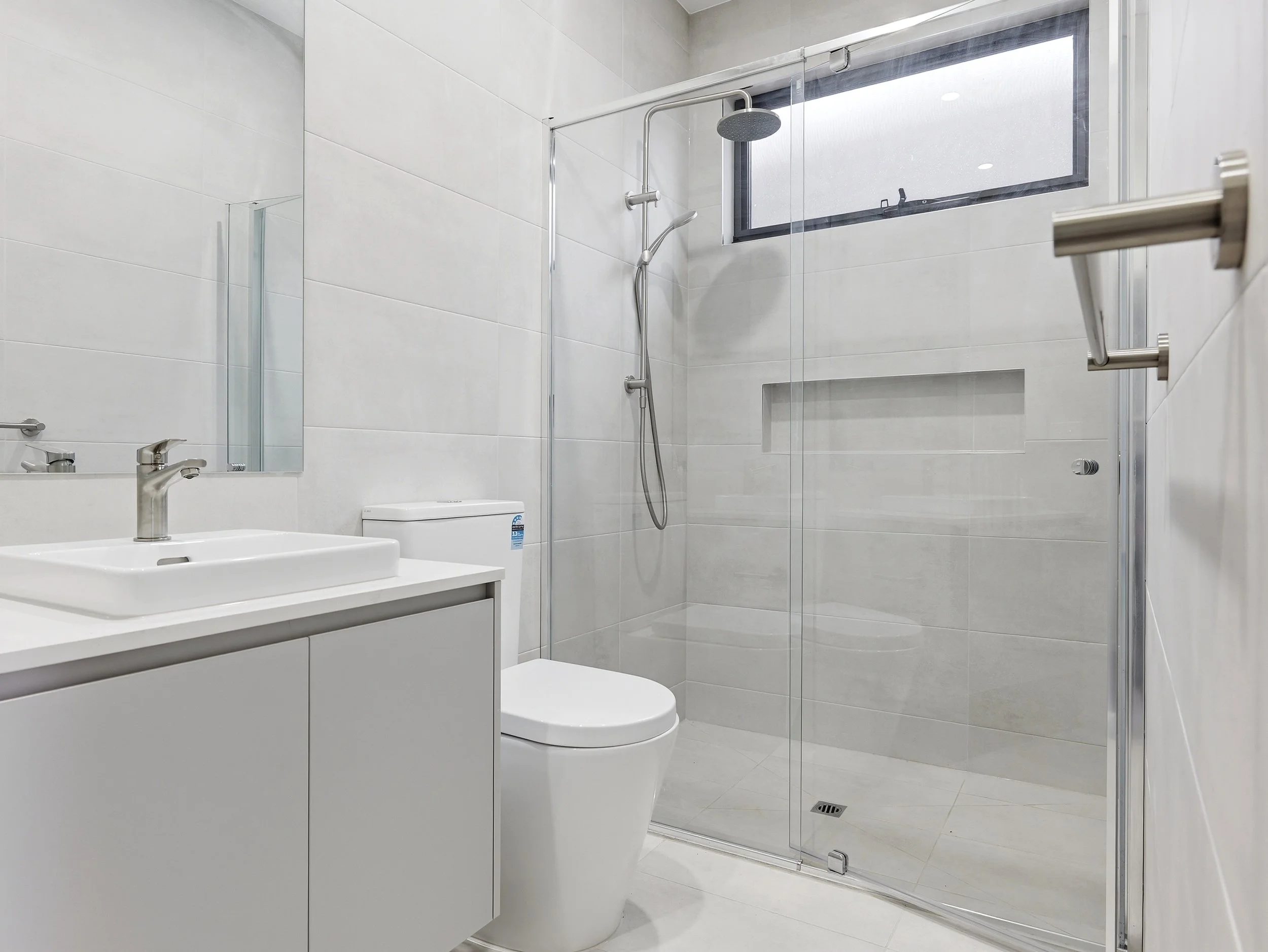 Modern bathroom with white tiles, a glass shower enclosure with a showerhead and a handheld shower, a white toilet, a sink with a mirror, and a small window.