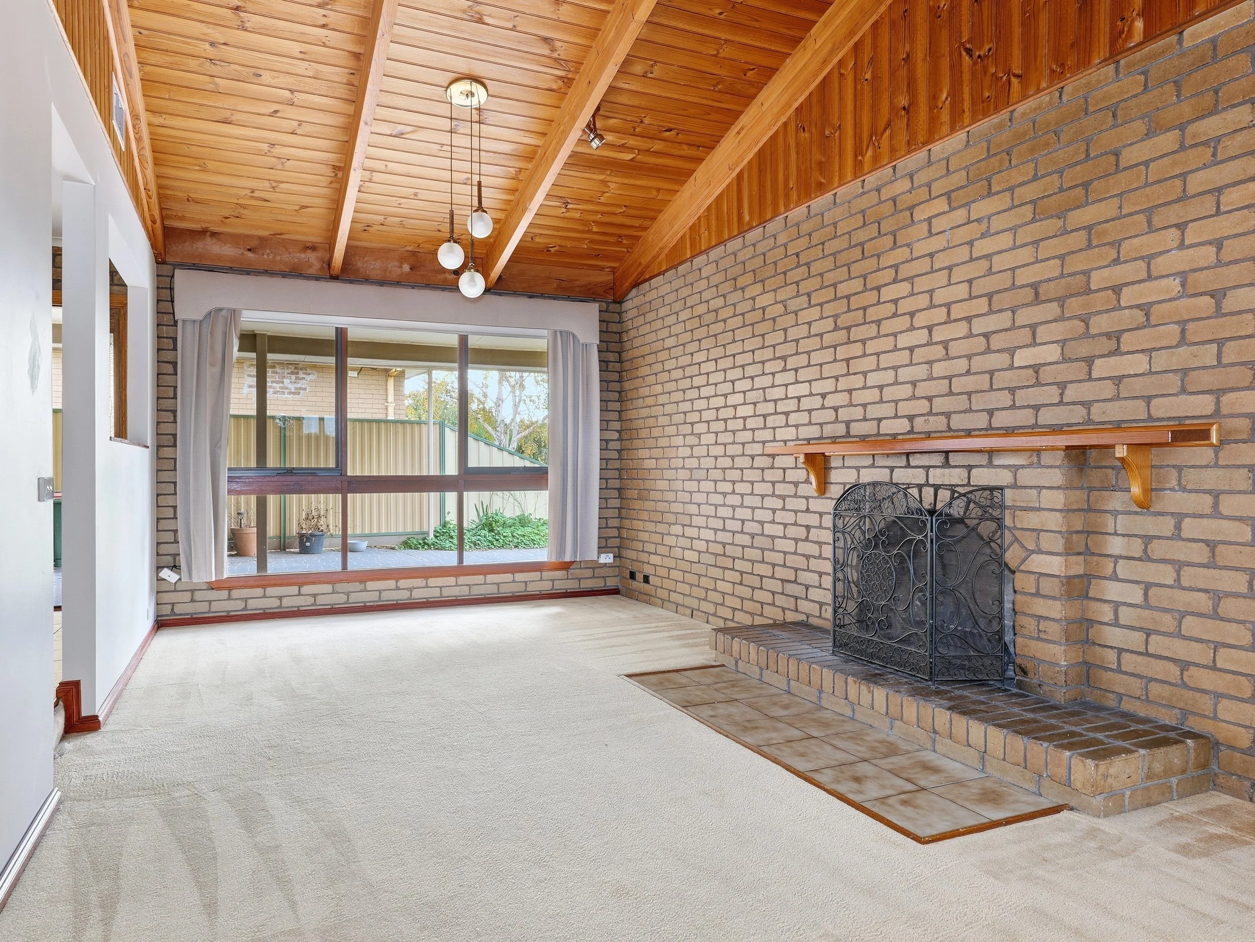 Living room with brick fireplace, carpeted floor, large window with curtains, and a wooden ceiling with pendant lights.